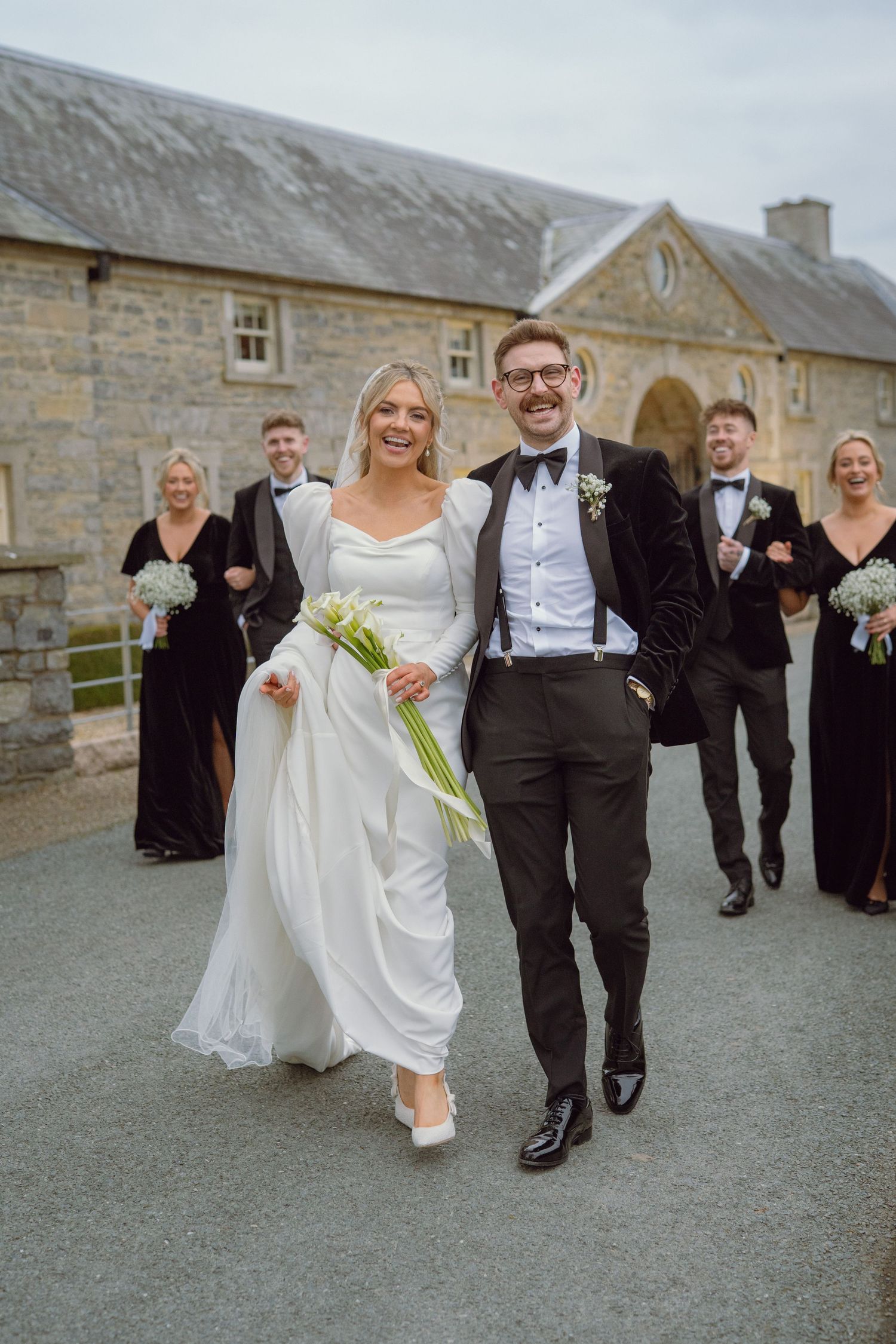 Smiling bride and groom leading wedding party outside a historic stone building.