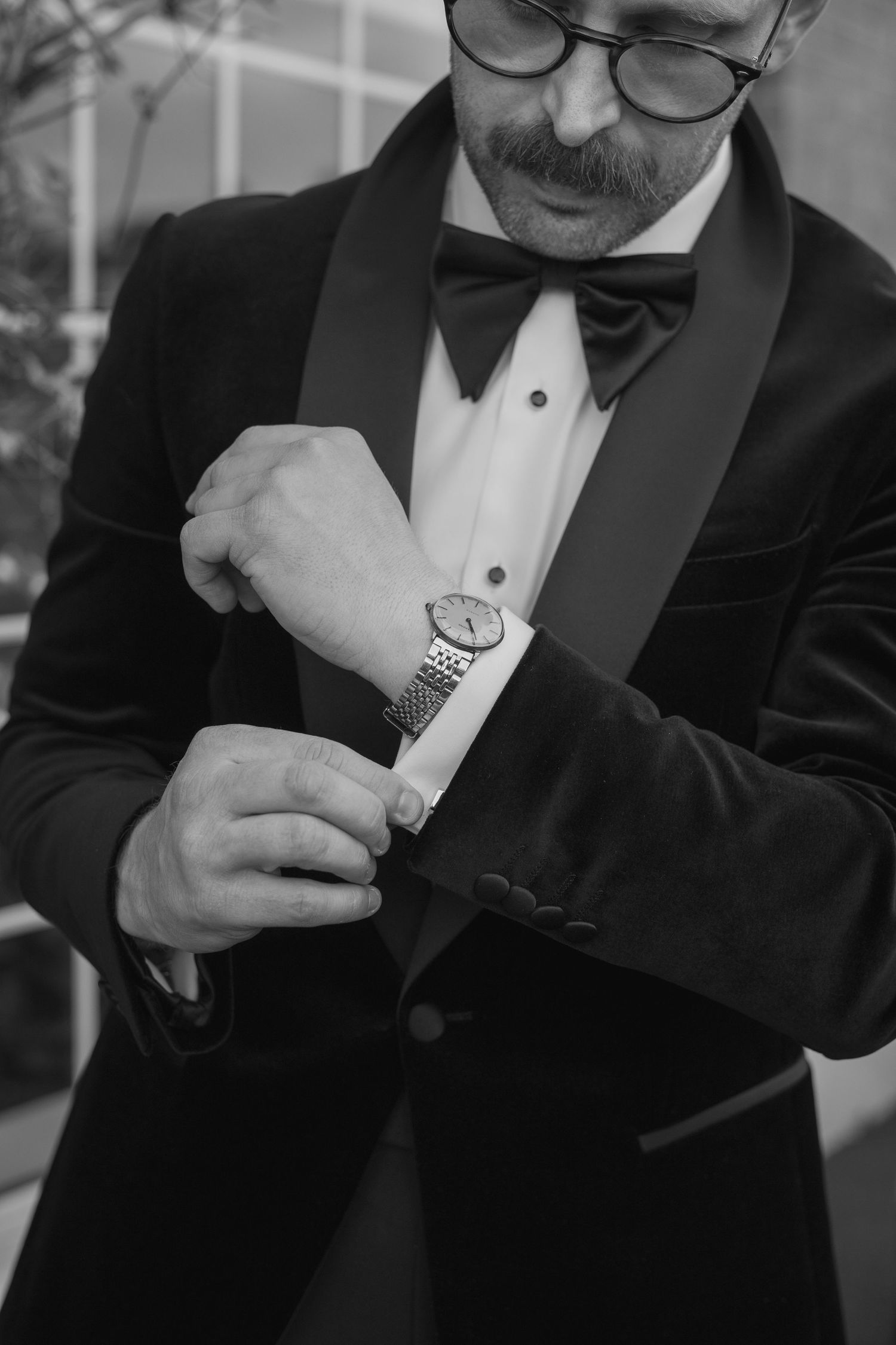 Dapper groom in black tuxedo and bow tie adjusting his watch before wedding.