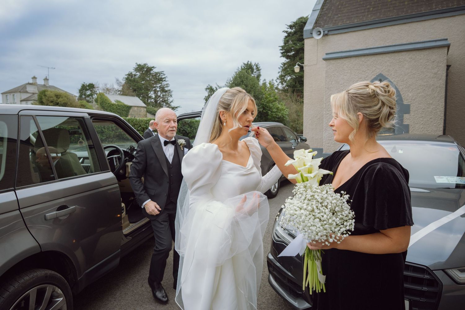 Bride in white gown and veil receiving bouquet from woman in black dress outside church.