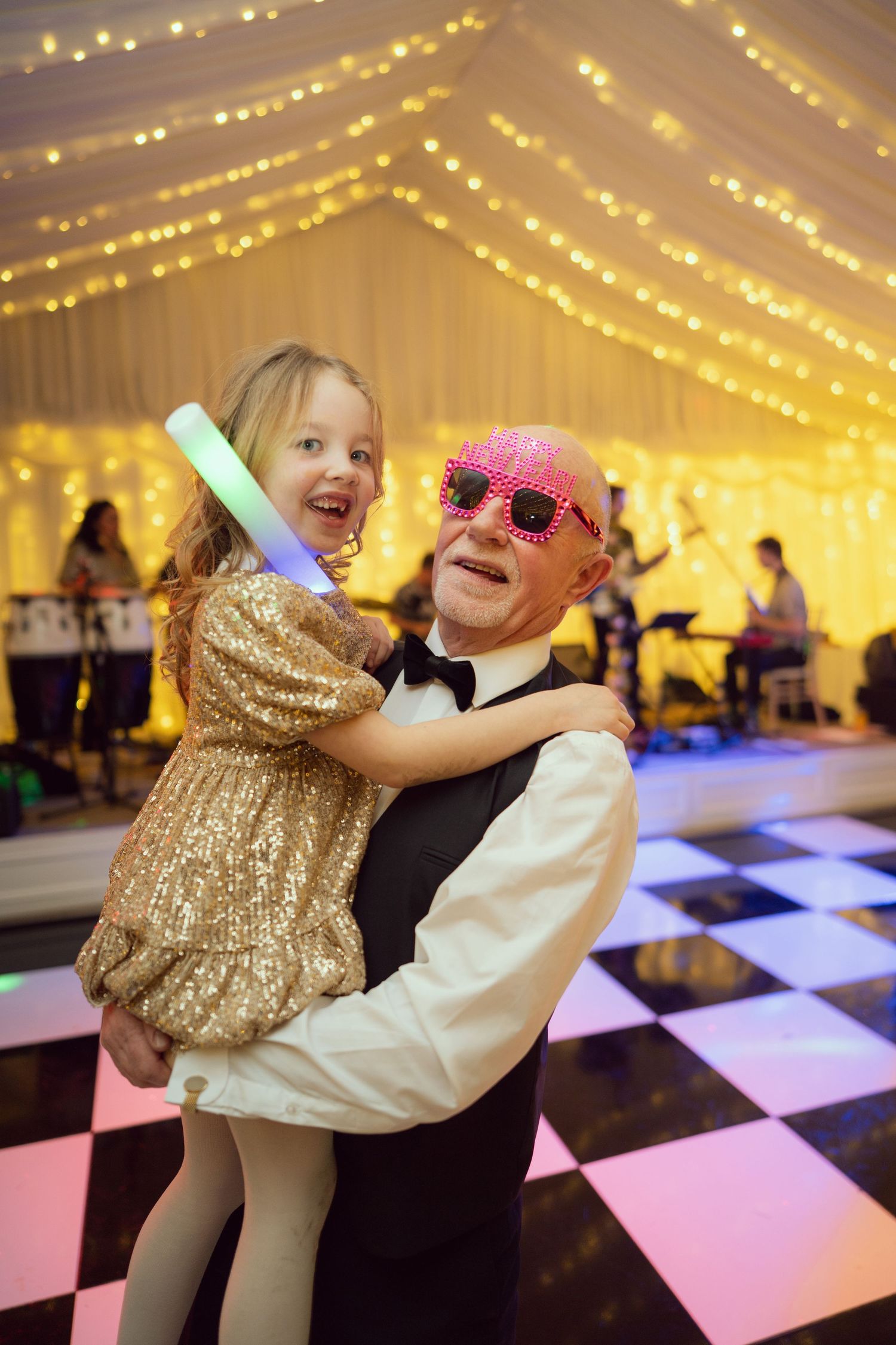 Elderly man wearing pink novelty sunglasses dancing with young girl in gold sequin dress at wedding reception.