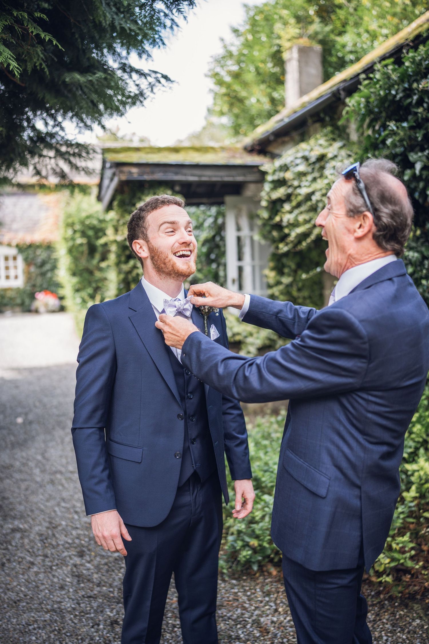 Groom laughing as older man adjusts his bow tie outside a rustic wedding venue.