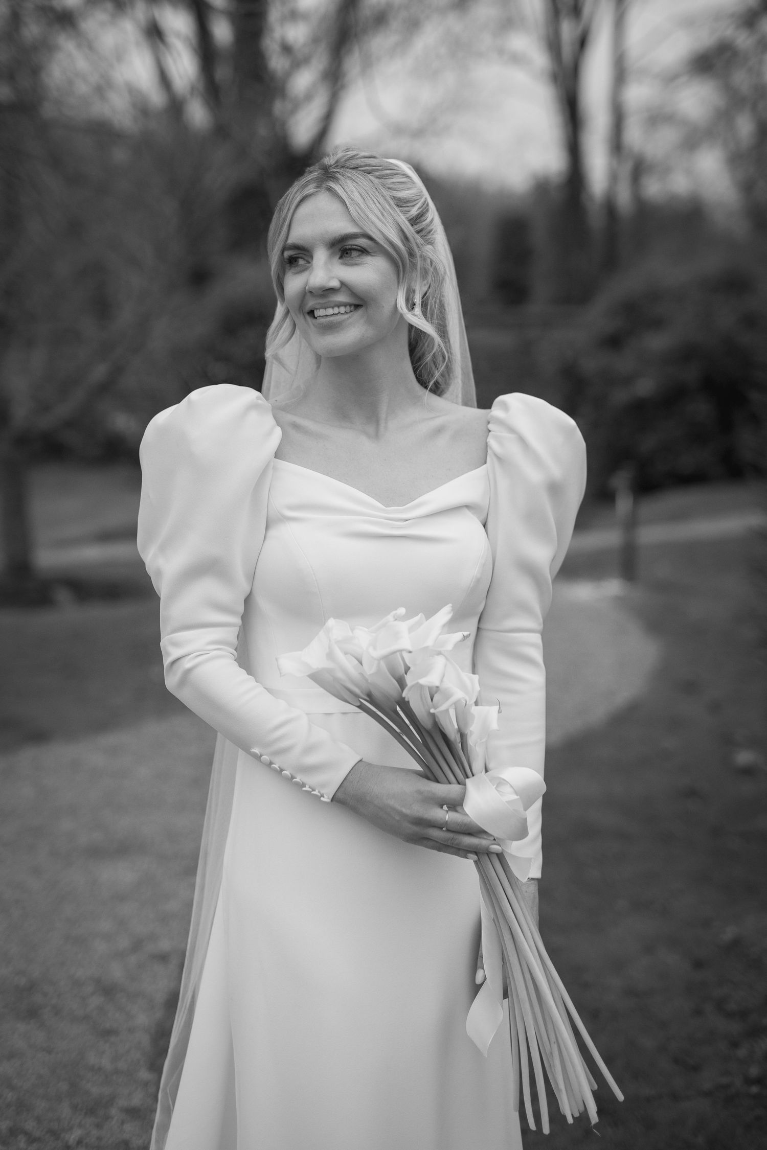 Smiling bride in elegant white puff sleeve wedding gown holding tulip bouquet outdoors in black and white photo.