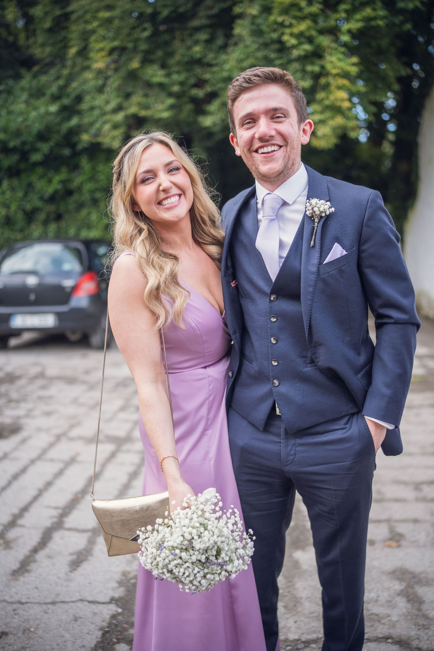 Smiling bridesmaid in purple dress holding baby's breath bouquet poses with groomsman in navy suit at wedding.