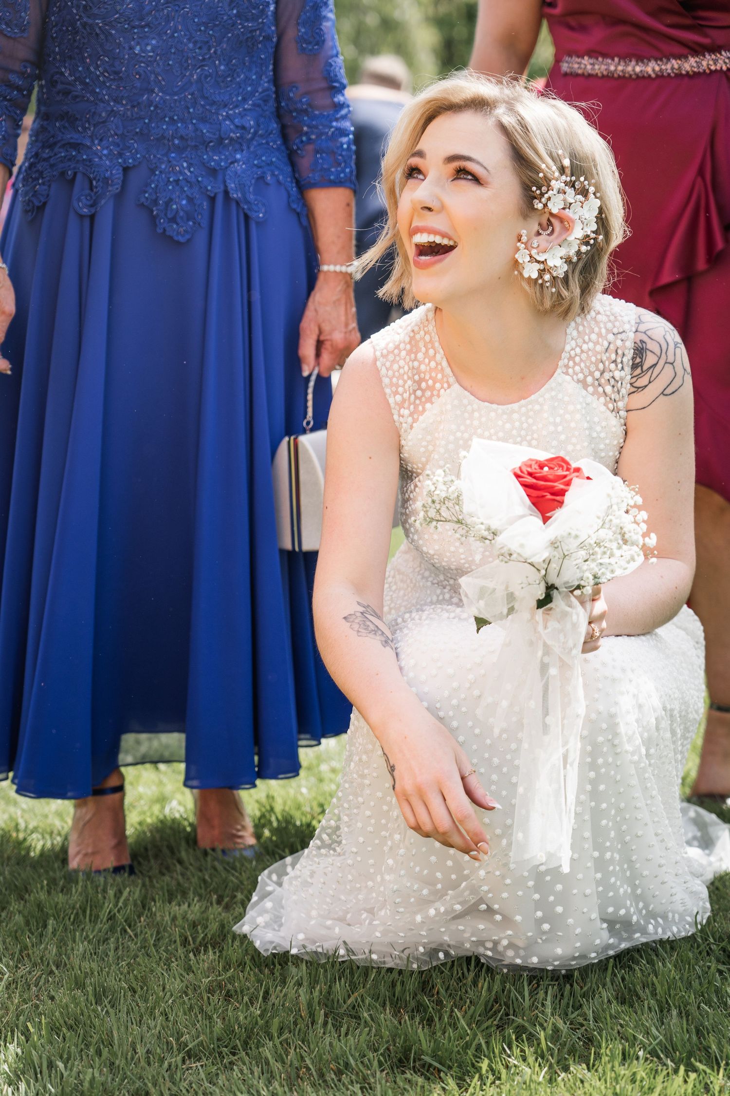 Smiling bride in polka-dot wedding dress kneeling on grass holding red and white bouquet at outdoor wedding.
