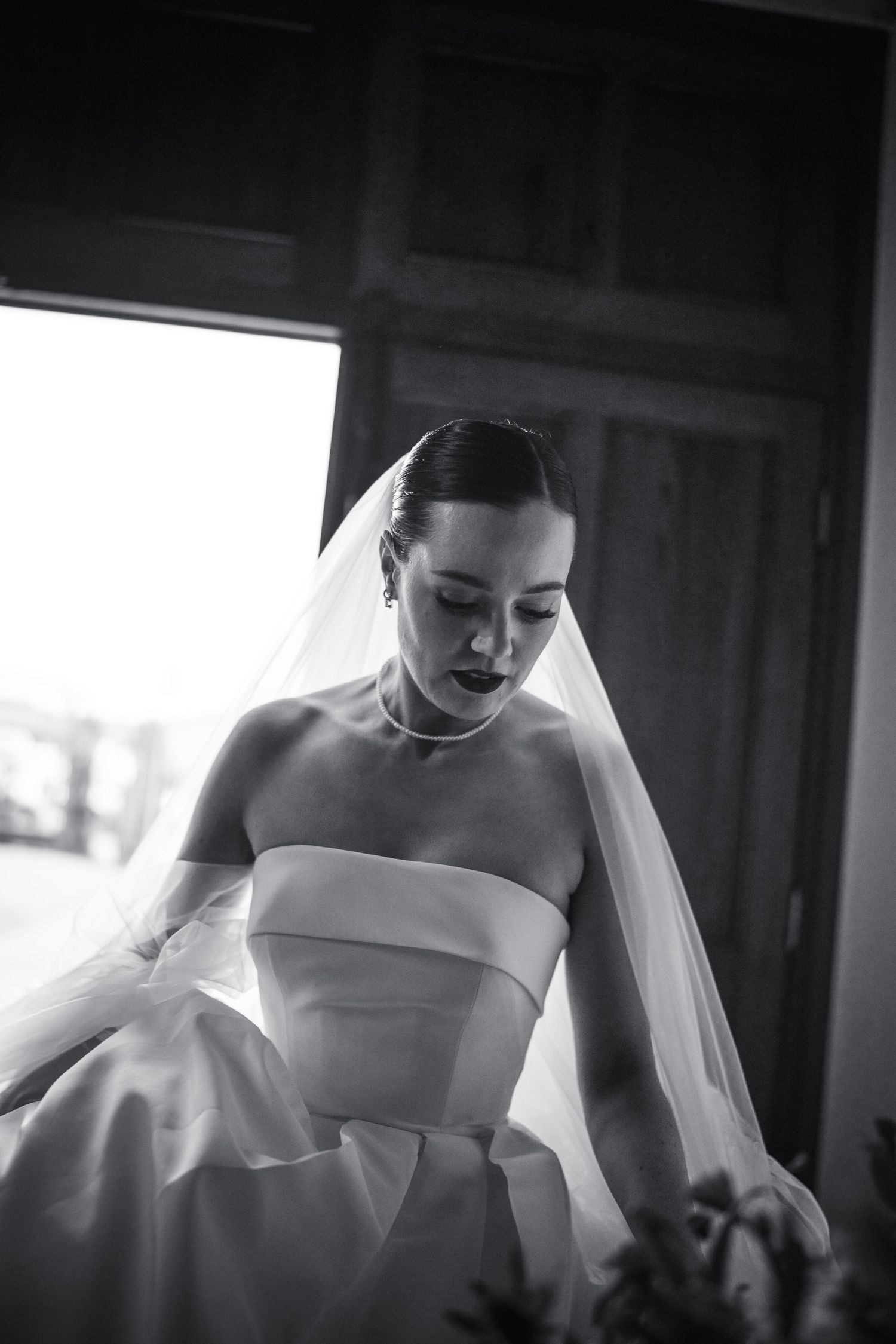 Bride in strapless white wedding gown and veil looking down, dramatic black and white photo.