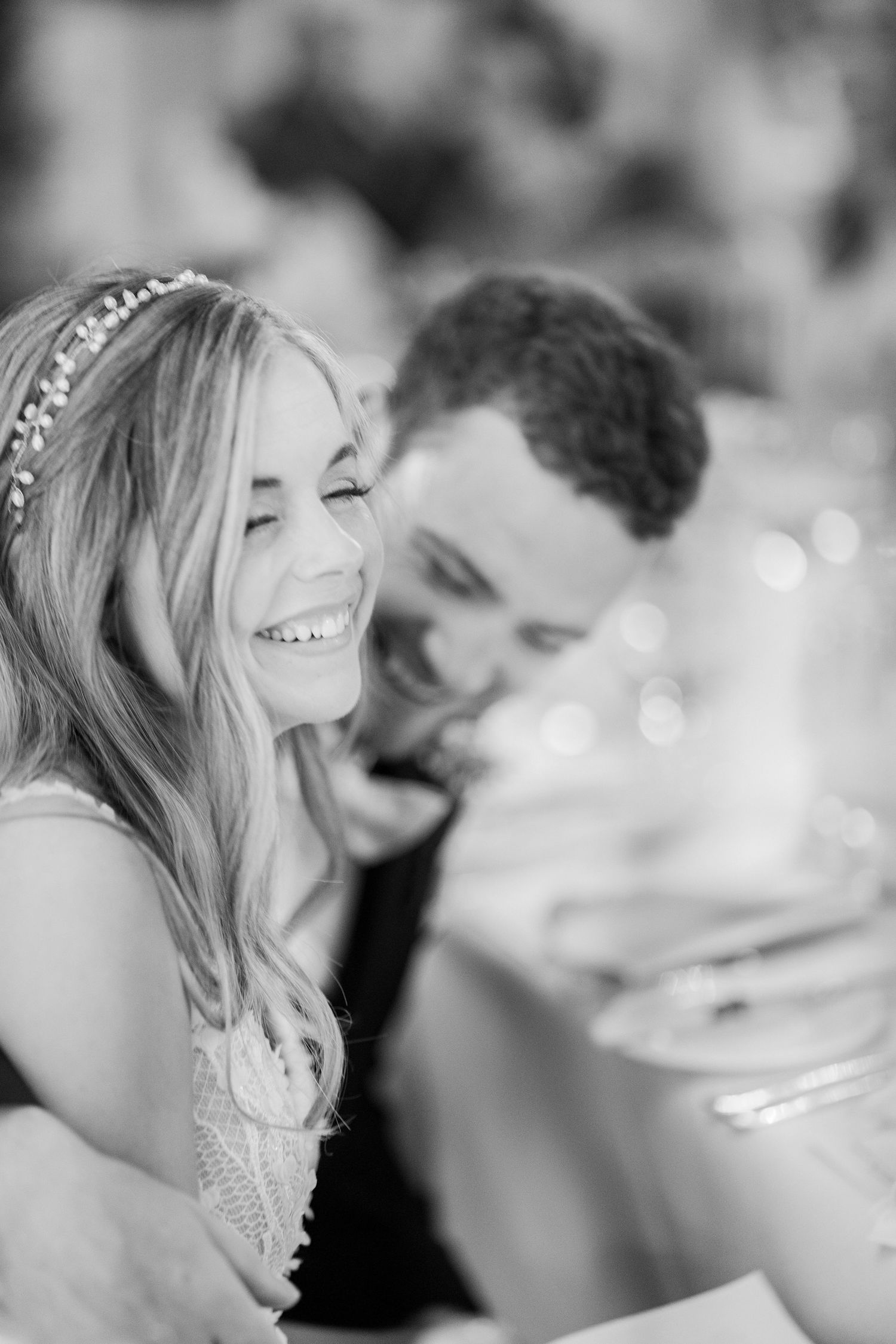 Black and white photo of smiling bride with floral headpiece and groom sharing a joyful moment at wedding reception.