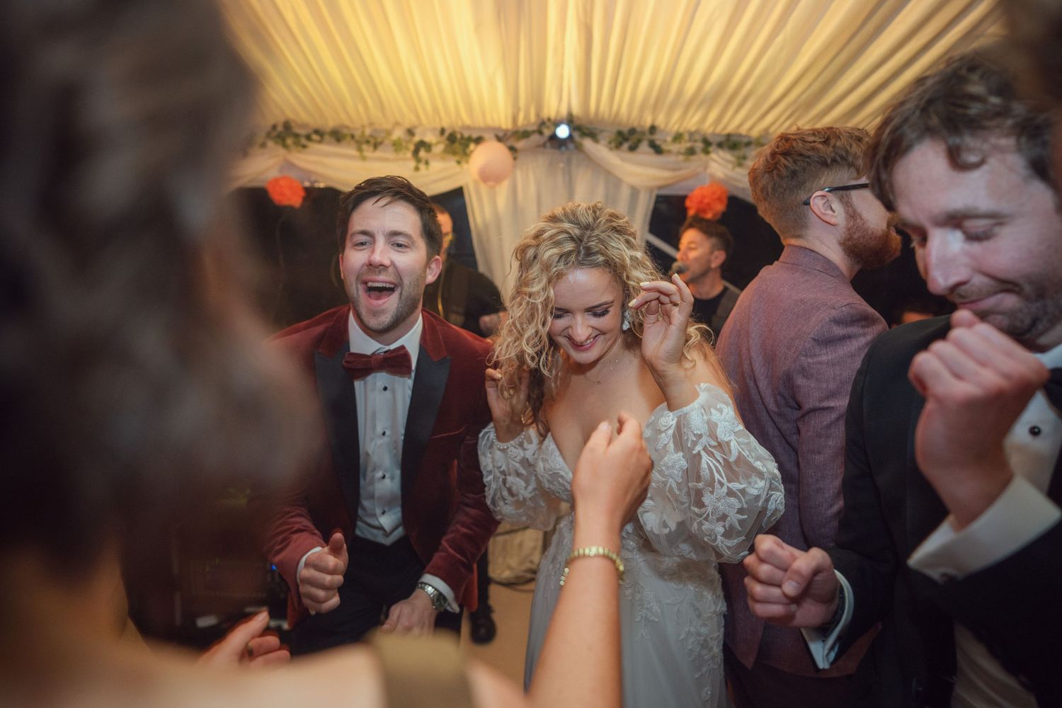 Bride in lace gown and groom in red velvet suit laugh joyfully on the wedding reception dance floor at garden wedding reception in Co Meath.