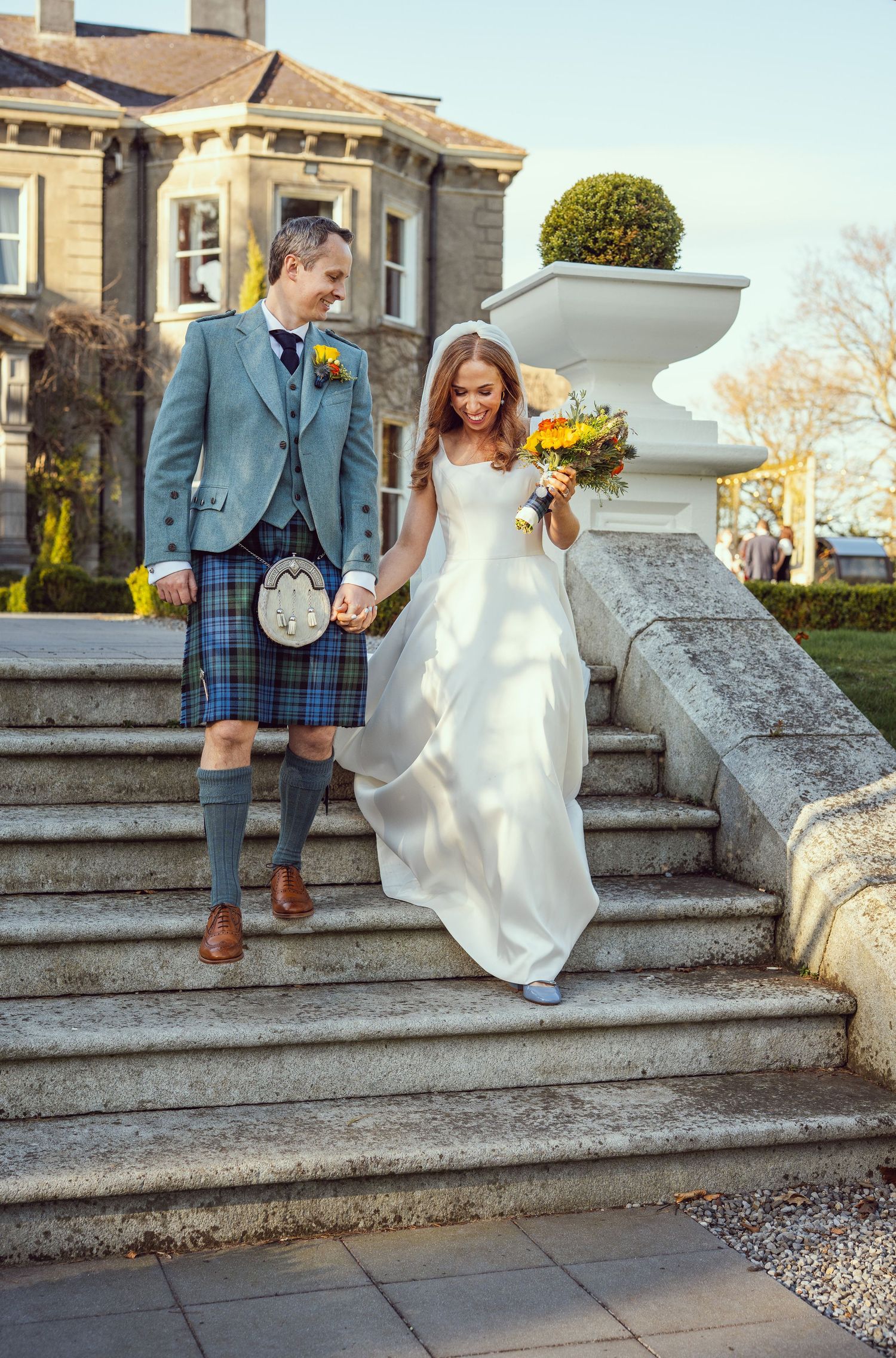 Bride and groom in blue tartan kilt descending grand stone steps at a Scottish country house wedding.
