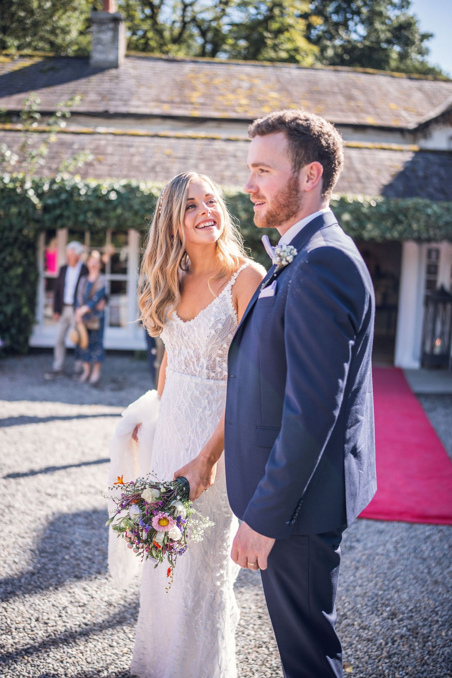 Smiling bride in lace gown holding wildflower bouquet stands with groom in navy suit outside venue.