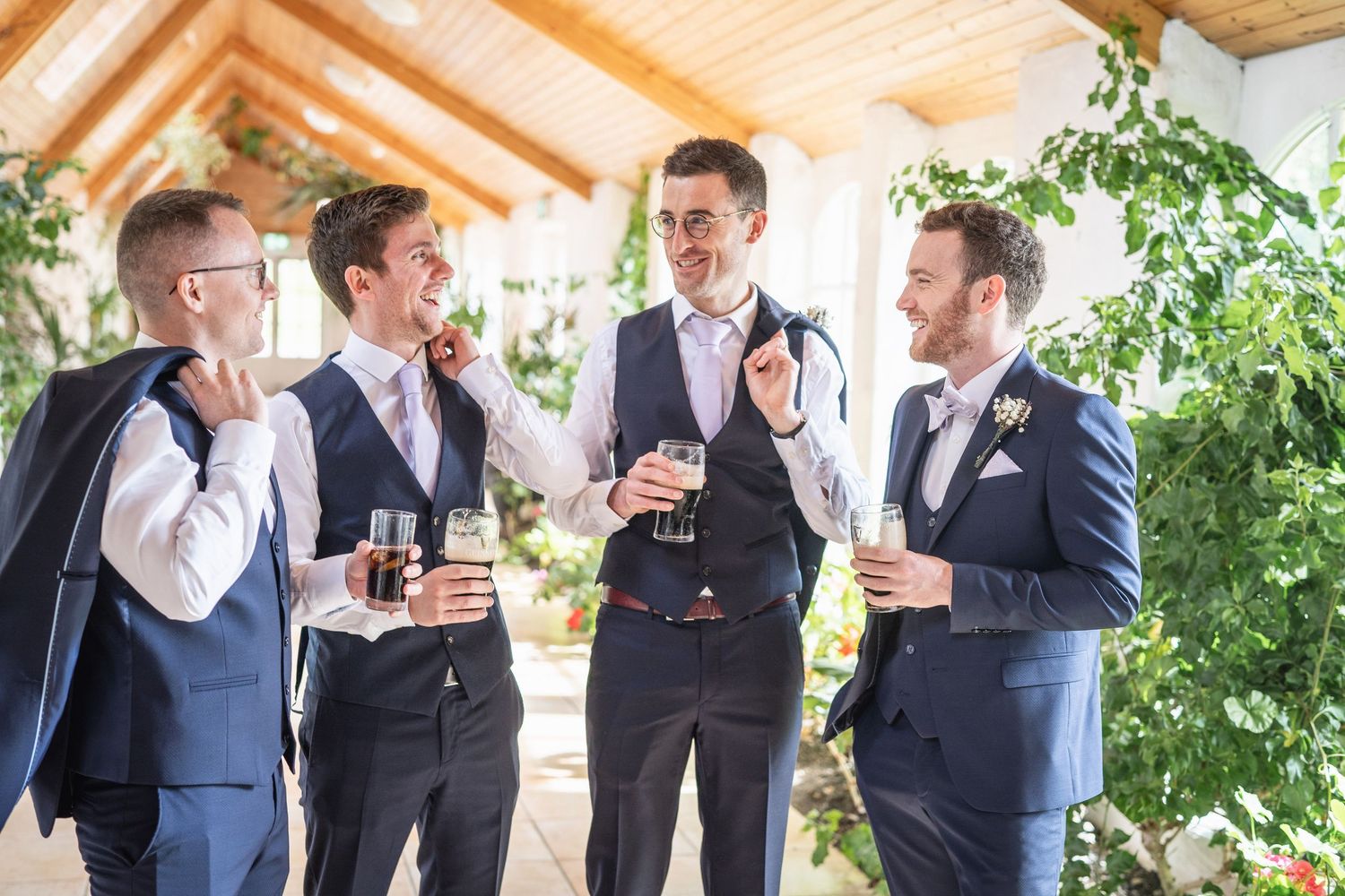 Four groomsmen in navy suits laugh and enjoy drinks together in a sunlit venue before the wedding ceremony.