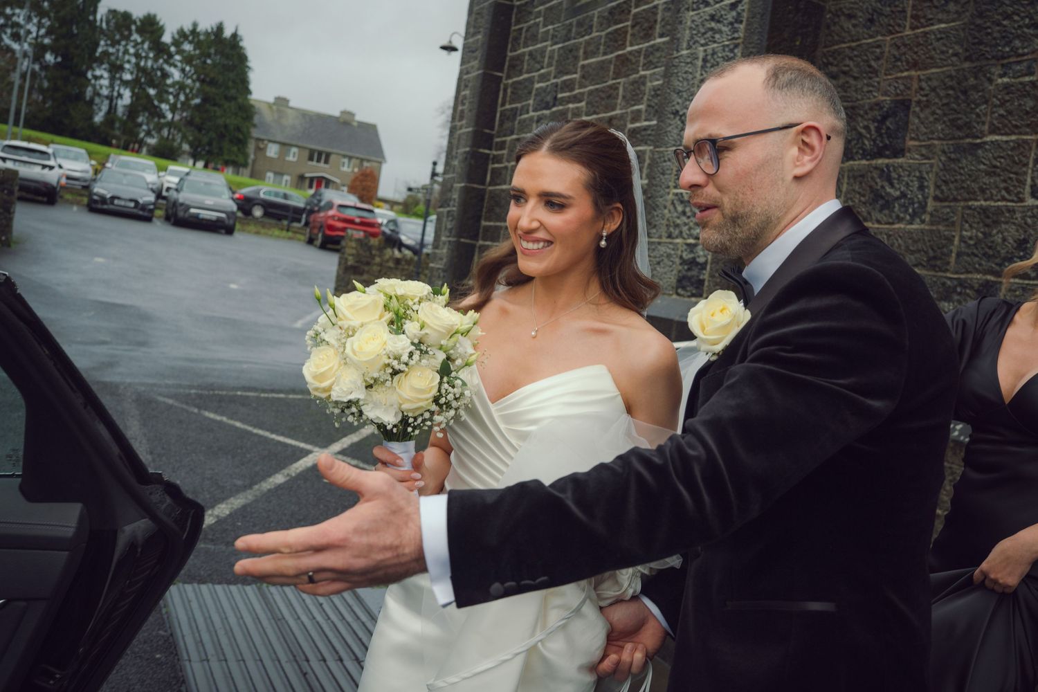 Smiling bride in strapless white dress holding bouquet with groom opening car door outside church at Mount Juliet wedding Kilkenny.