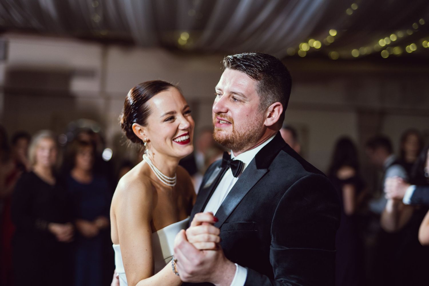 Bride and groom sharing first dance, bride in strapless white gown with pearl jewelry, groom in classic black tuxedo.