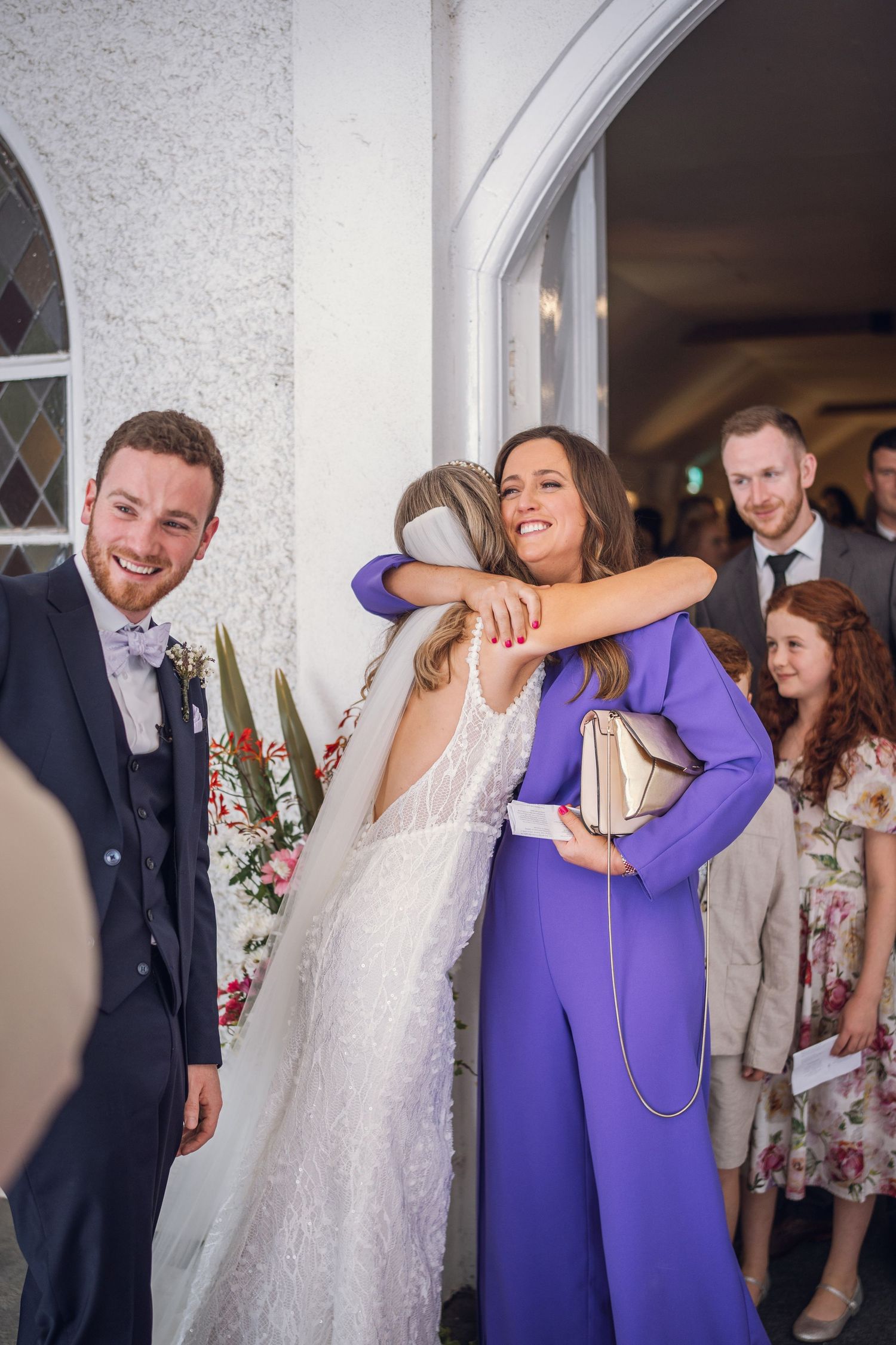 Groom smiles as bridesmaid in purple jumpsuit warmly hugs bride outside white church after ceremony.