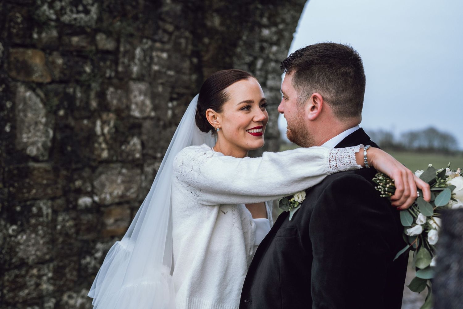 Smiling bride in white gown and veil embraces groom in black suit near stone wall.