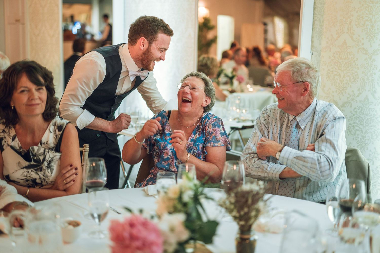 Groom in waistcoat sharing a laugh with elderly wedding guests seated at flower-decorated reception table.
