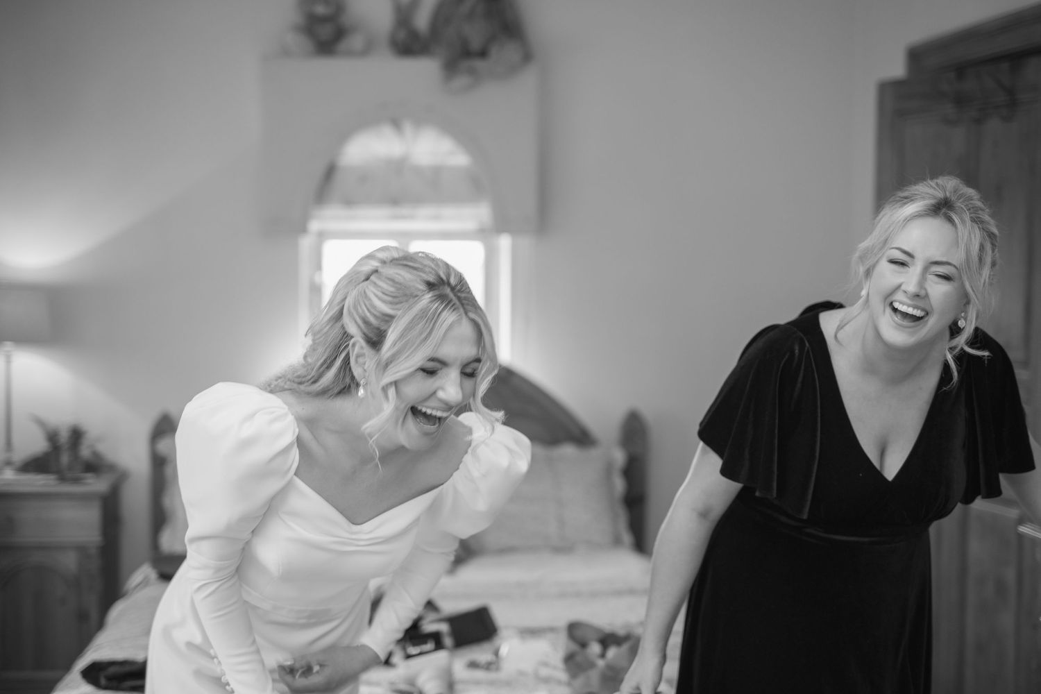 Bride and bridesmaid laugh joyfully together in black and white candid getting-ready photo.