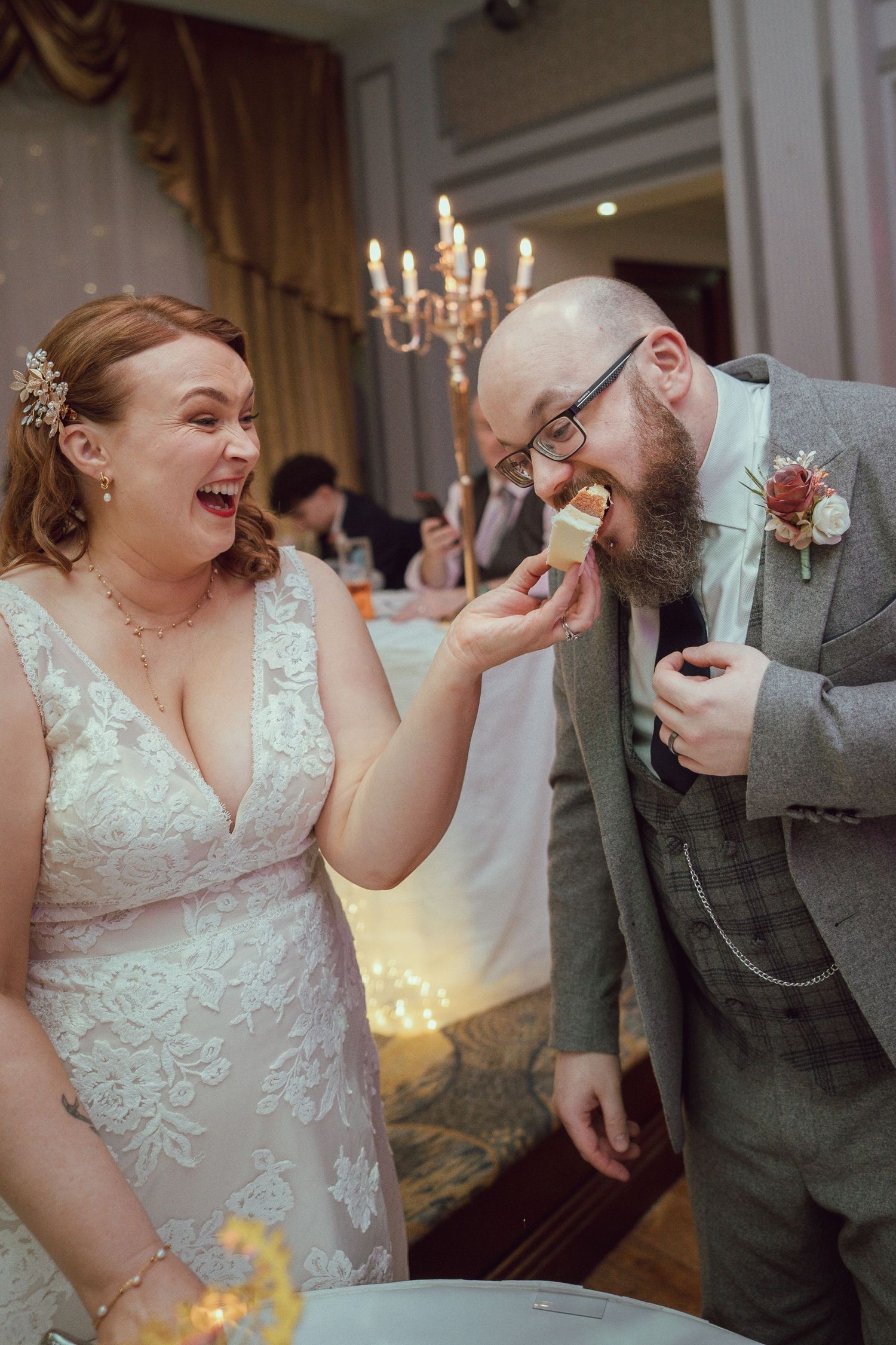 Joyful bride in lace dress feeding wedding cake to bearded groom in grey suit at elegant reception venue.