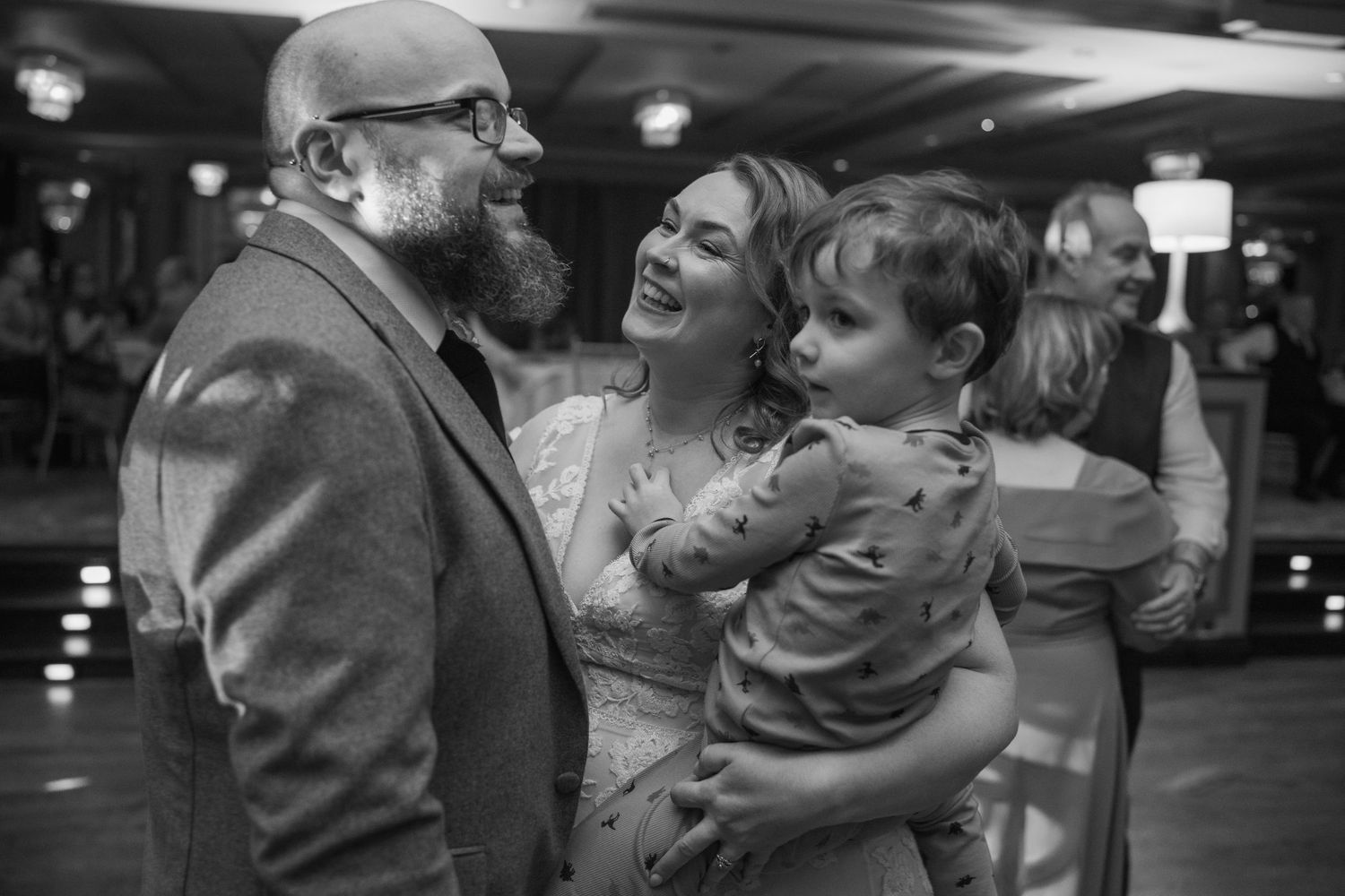 Black and white candid photo of bearded groom and laughing bride holding a young child on the dance floor.