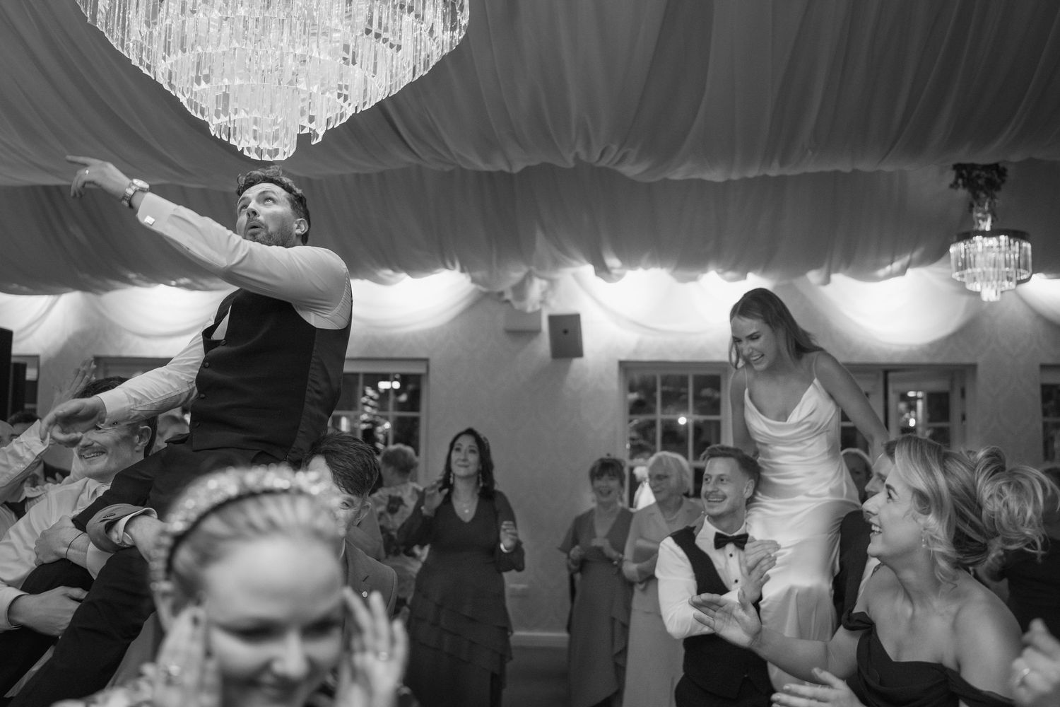Bride and groom lifted high on shoulders by guests during energetic wedding celebration.