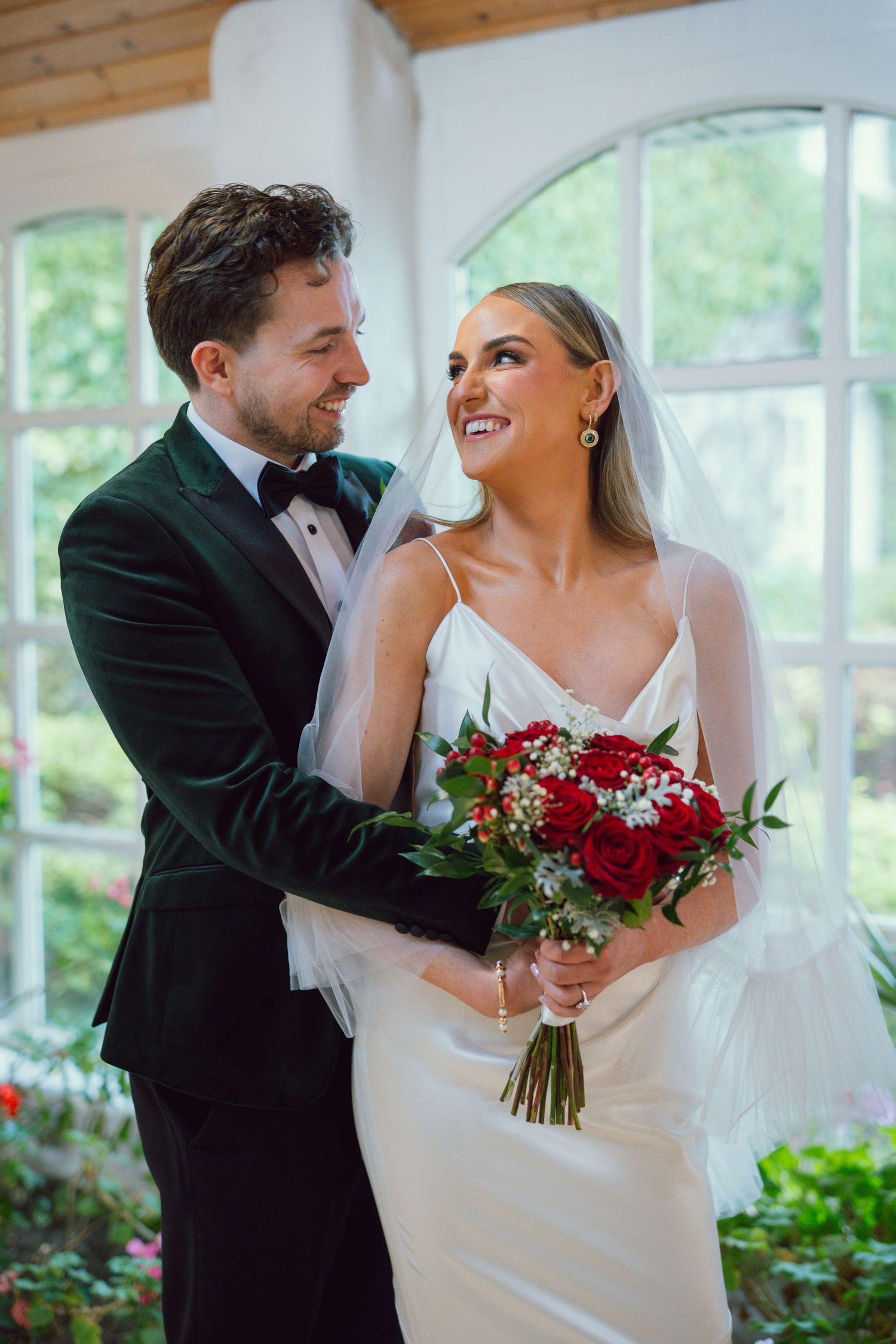 Bride and groom embracing in greenhouse holding red rose bouquet.