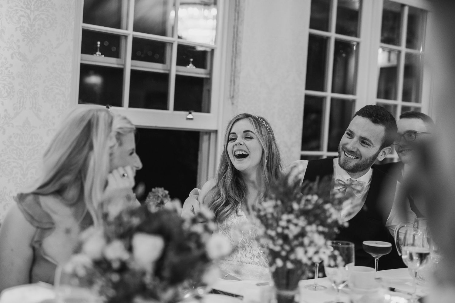 Black and white photo of laughing bride and groom enjoying reception dinner with guests at table.