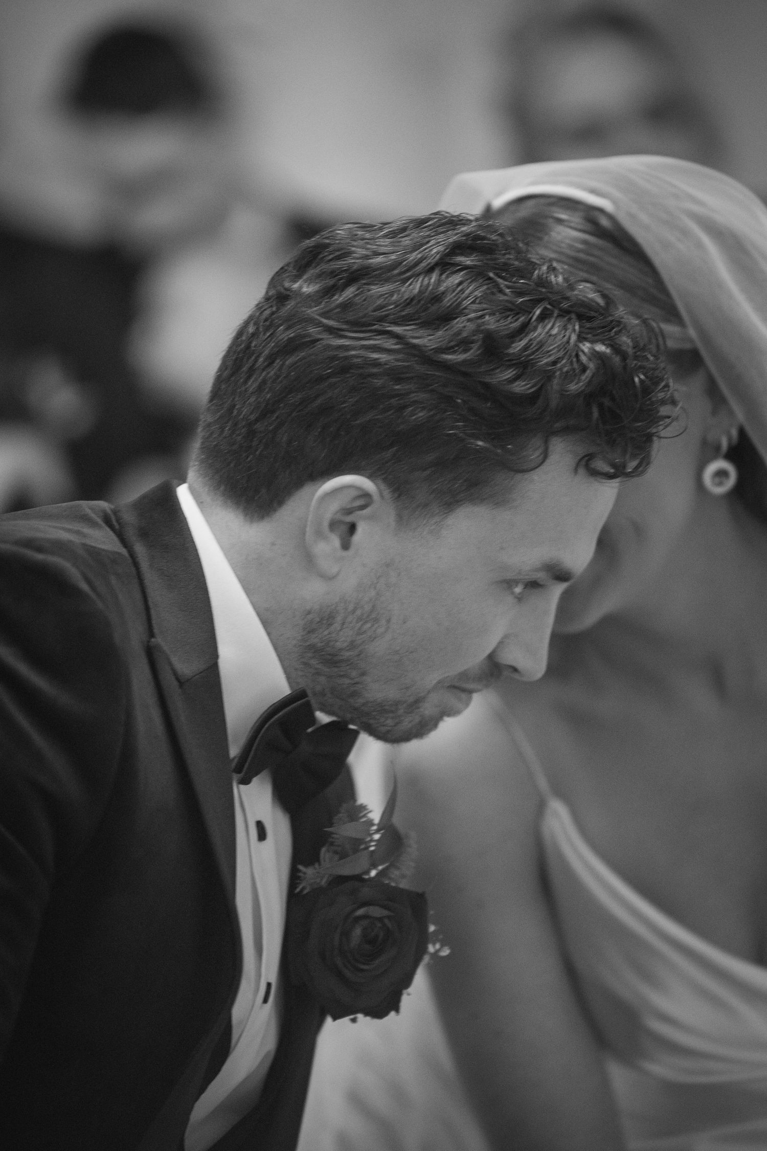 Groom in tuxedo with rose boutonniere bows head beside bride in veil during wedding ceremony.