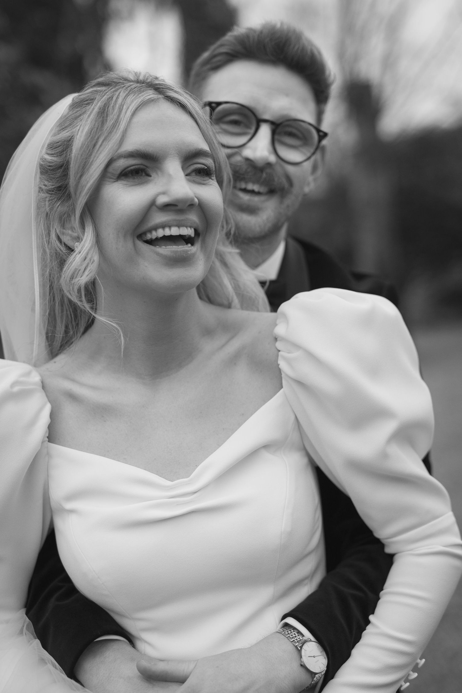 Laughing bride in white puff sleeve gown with groom wearing glasses embracing outdoors in black and white.