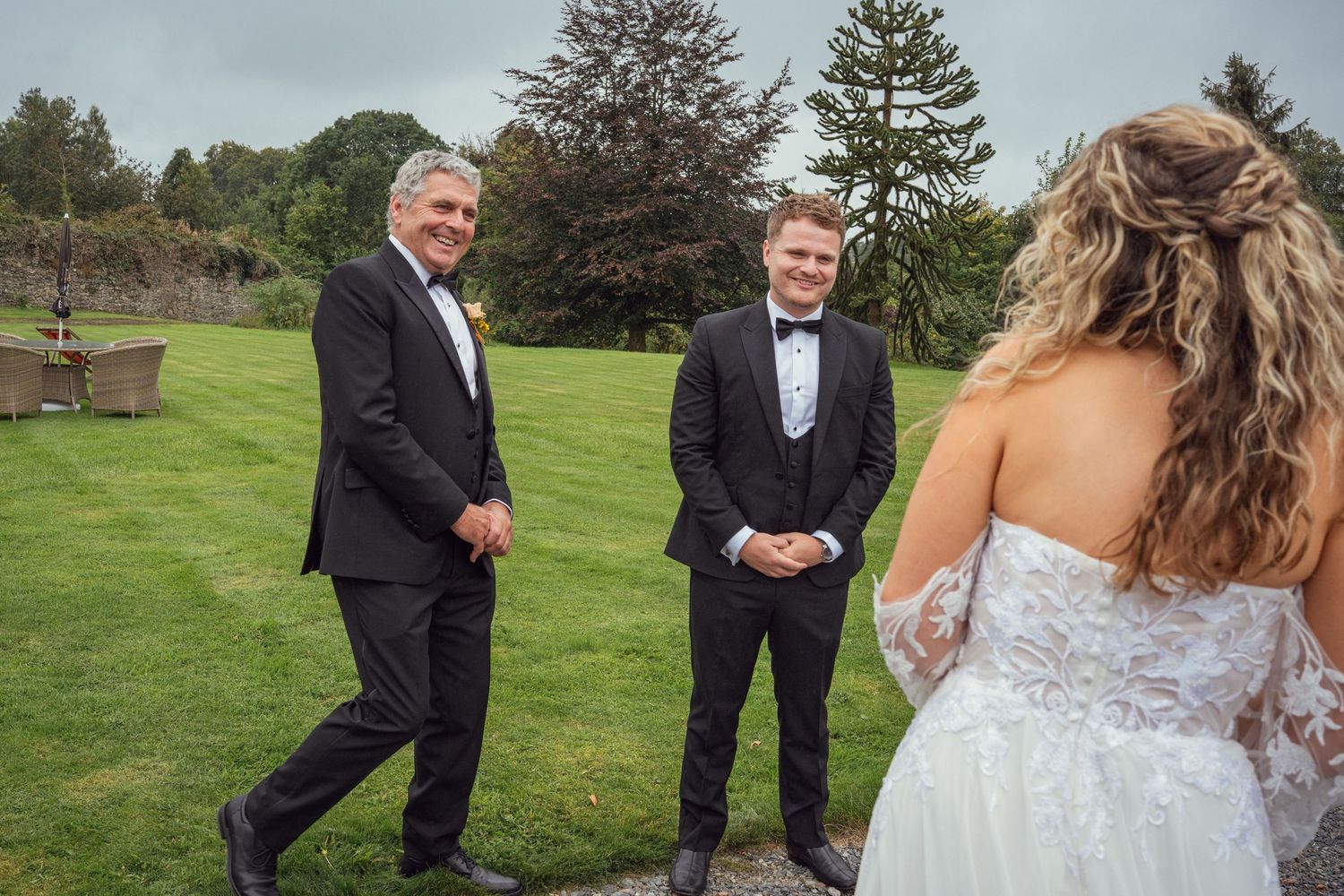 Groom smiling as he sees his bride approaching across a lush green lawn with her father.