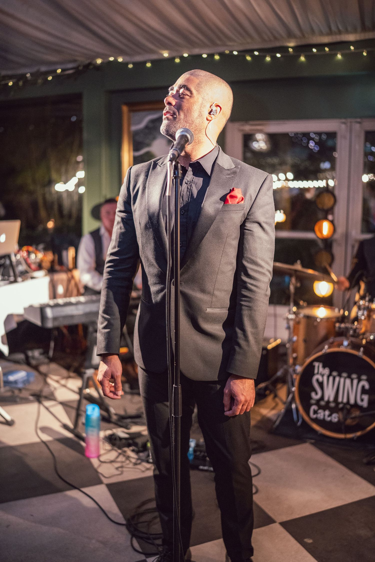 Bald male vocalist in grey suit performing passionately on stage for The Swing Cats band at a wedding reception.