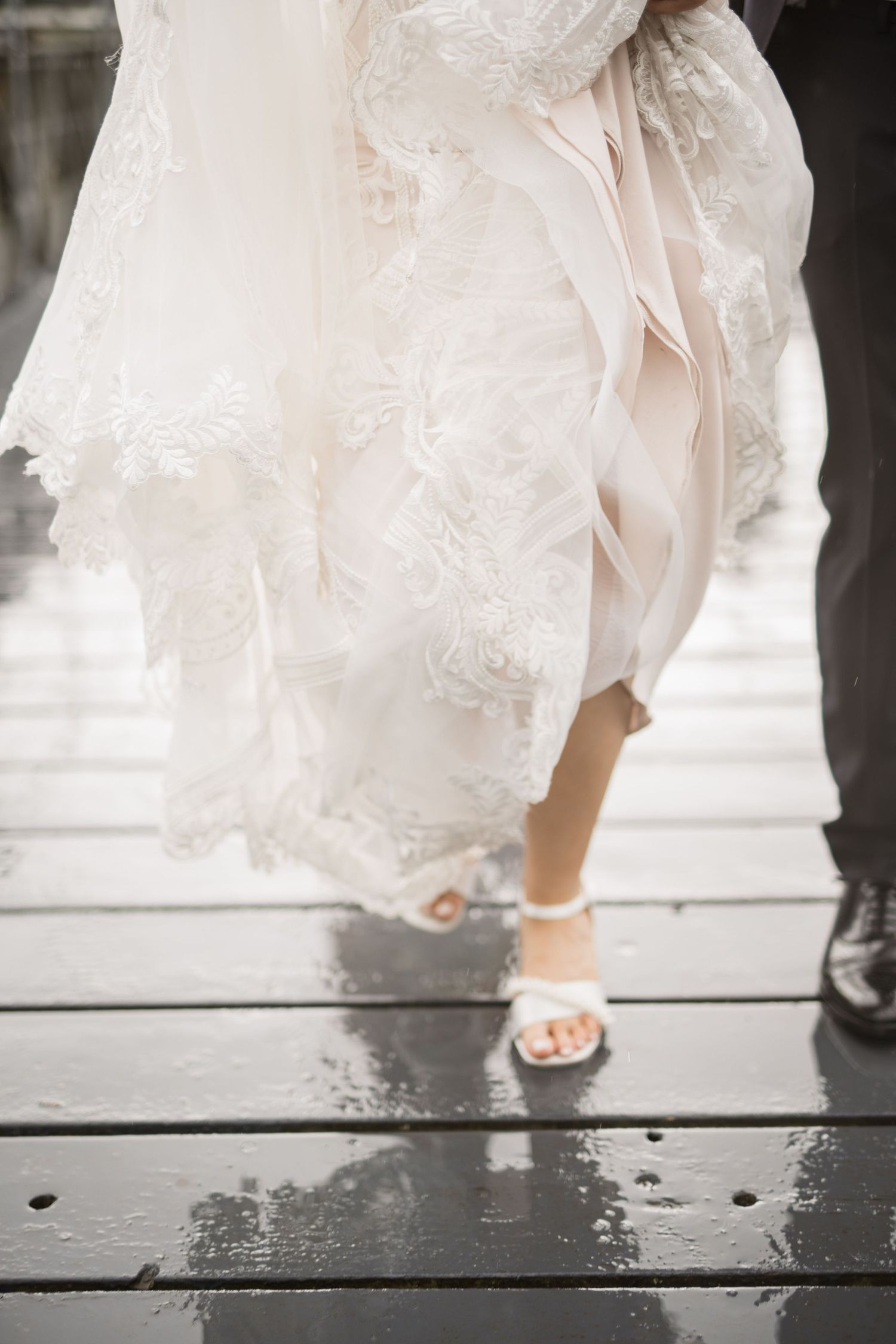 Close-up of bride's lace wedding gown hem and white strappy heels on wet wooden surface in rain.