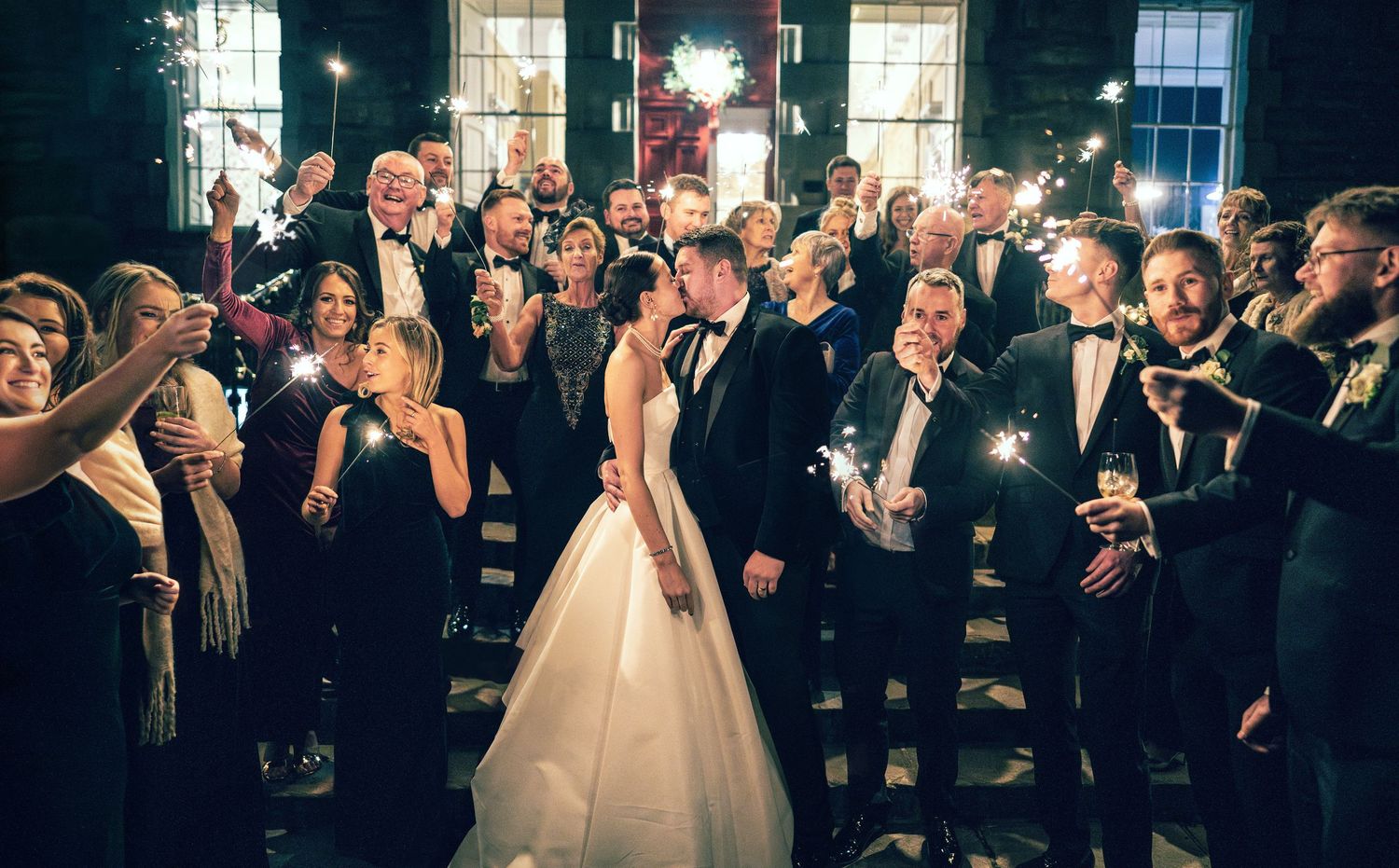 Bride and groom share a kiss surrounded by guests holding sparklers in a romantic nighttime celebration.