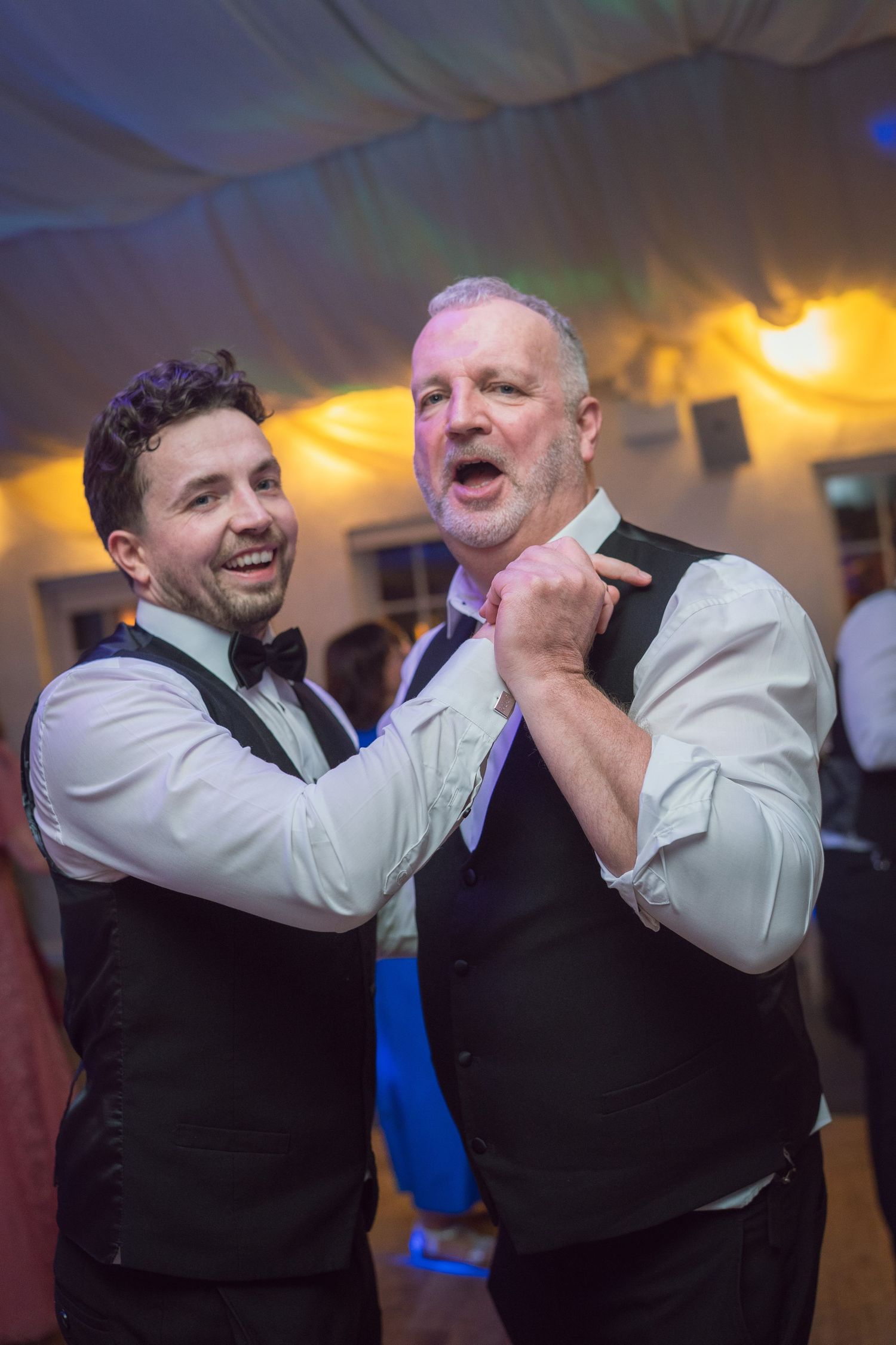 Two men in black vests and white shirts dancing joyfully at a wedding reception celebration.