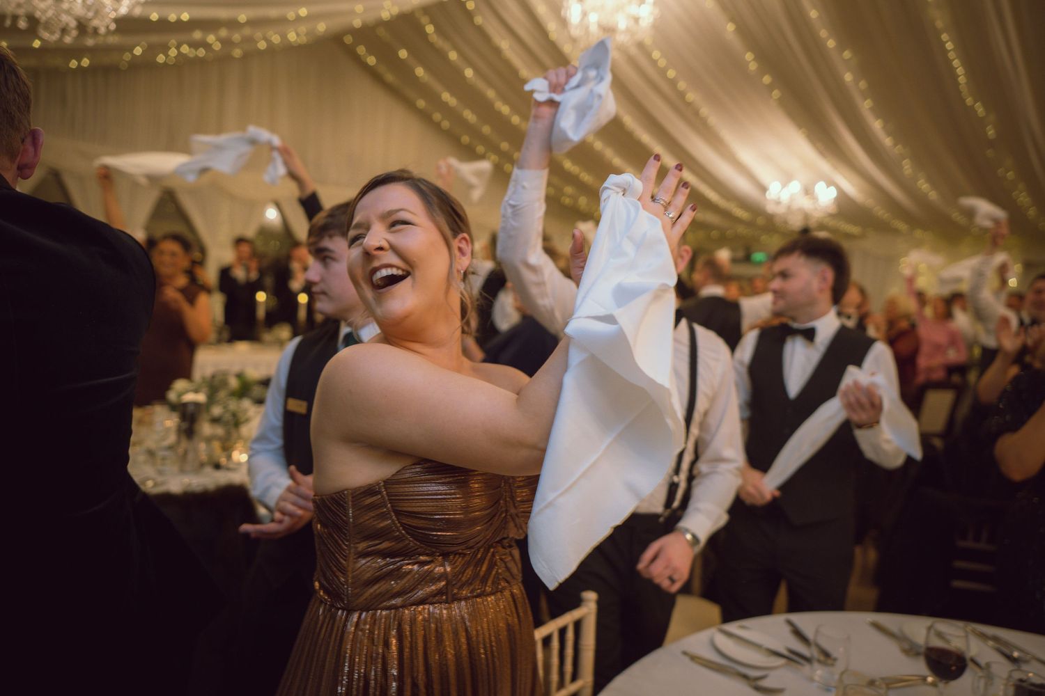 Joyful wedding guest in gold dress waving white napkin during lively reception celebration.