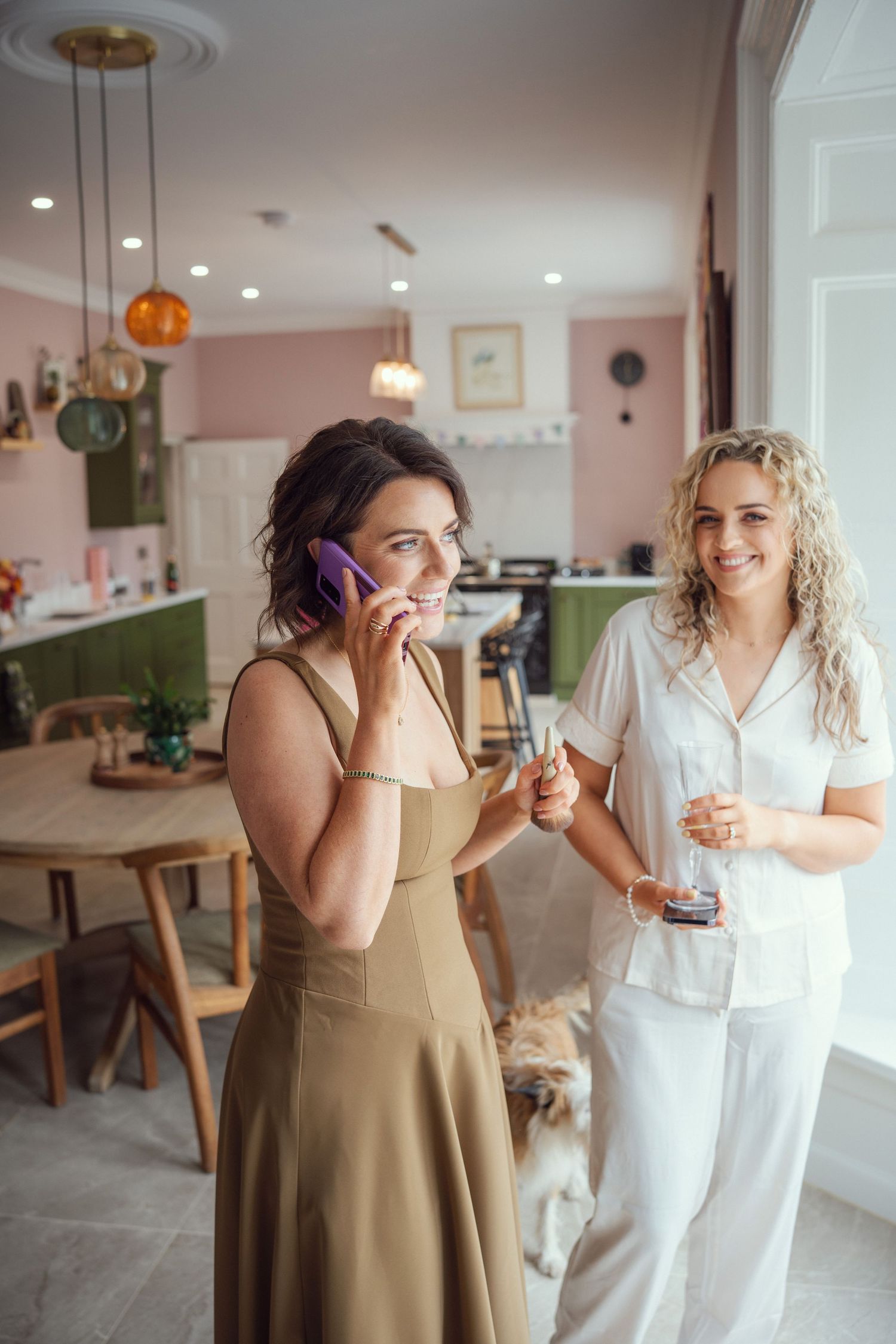 Bridesmaid in olive dress talks on purple phone while blonde woman in white pajamas smiles beside her.