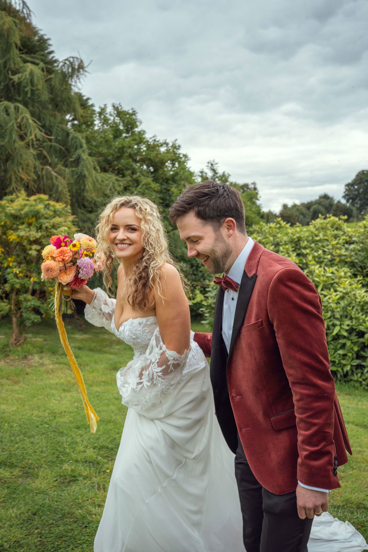 Smiling bride holding vibrant bouquet beside groom in burgundy velvet jacket outdoors outdoor garden wedding reception in Co Meath.