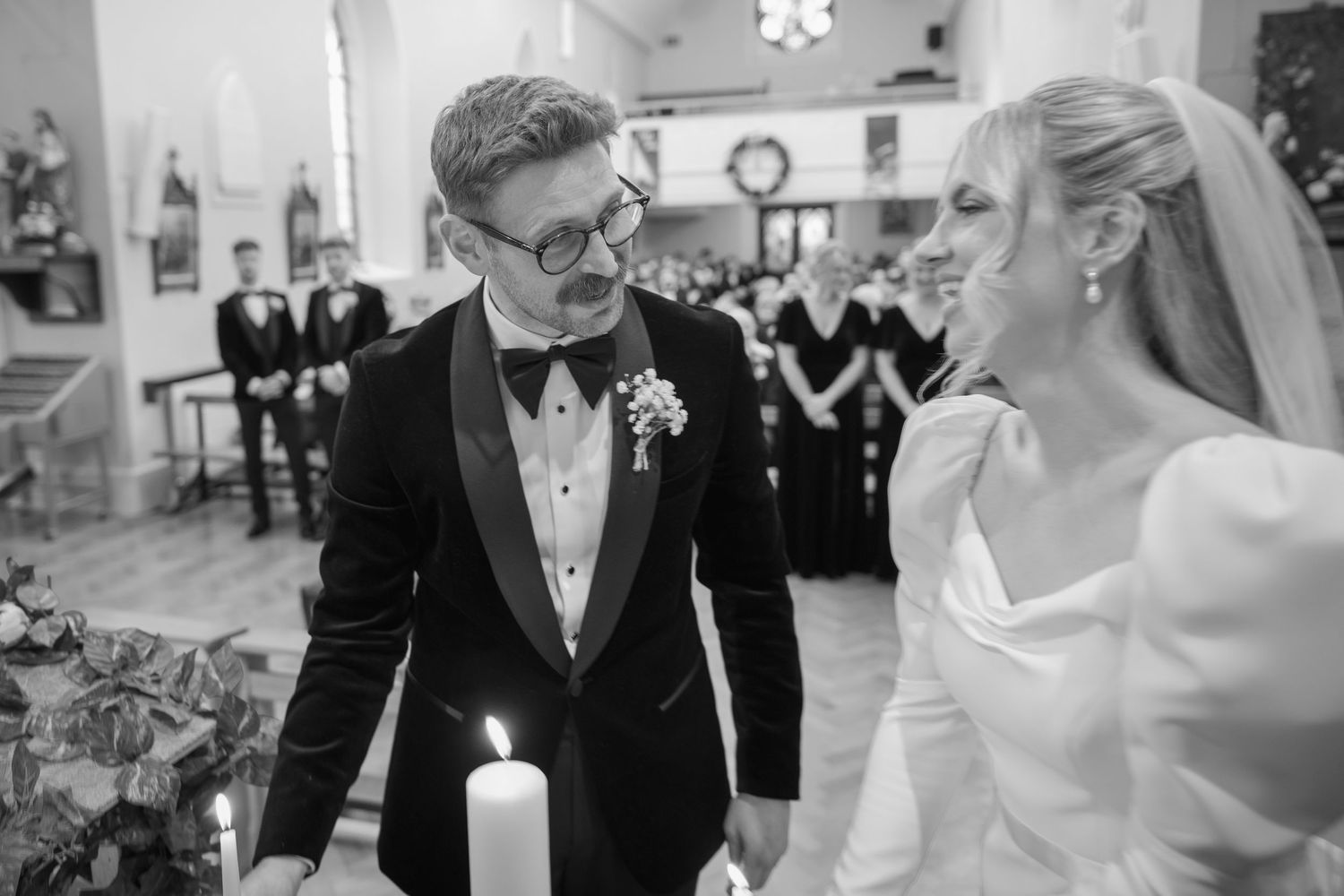 Black and white photo of newlyweds sharing a smile by candlelight at church altar.