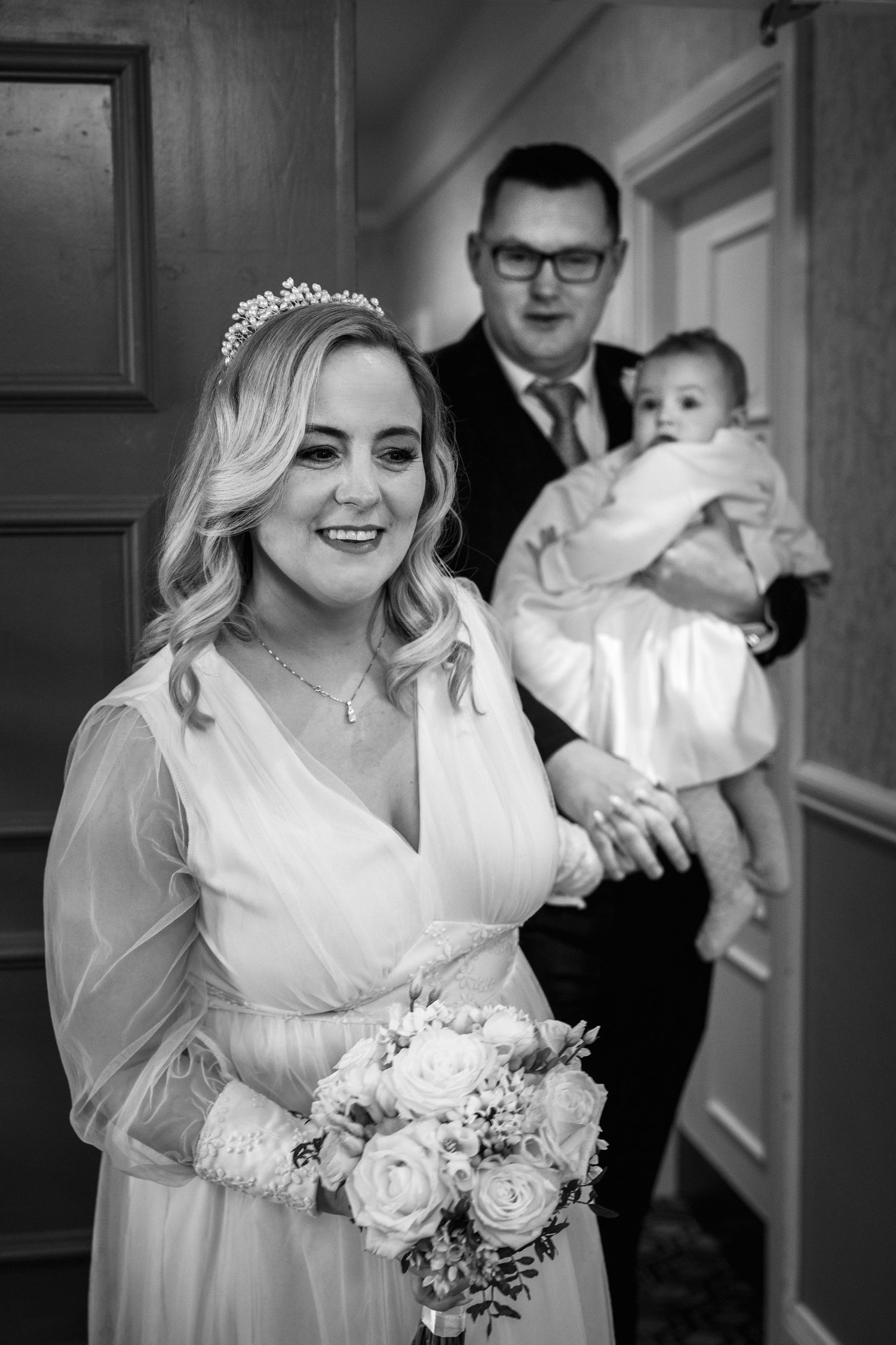 Bride in white gown holding rose bouquet smiles as groom holds baby behind her at Headfort Arm Hotel wedding in Co. Meath.