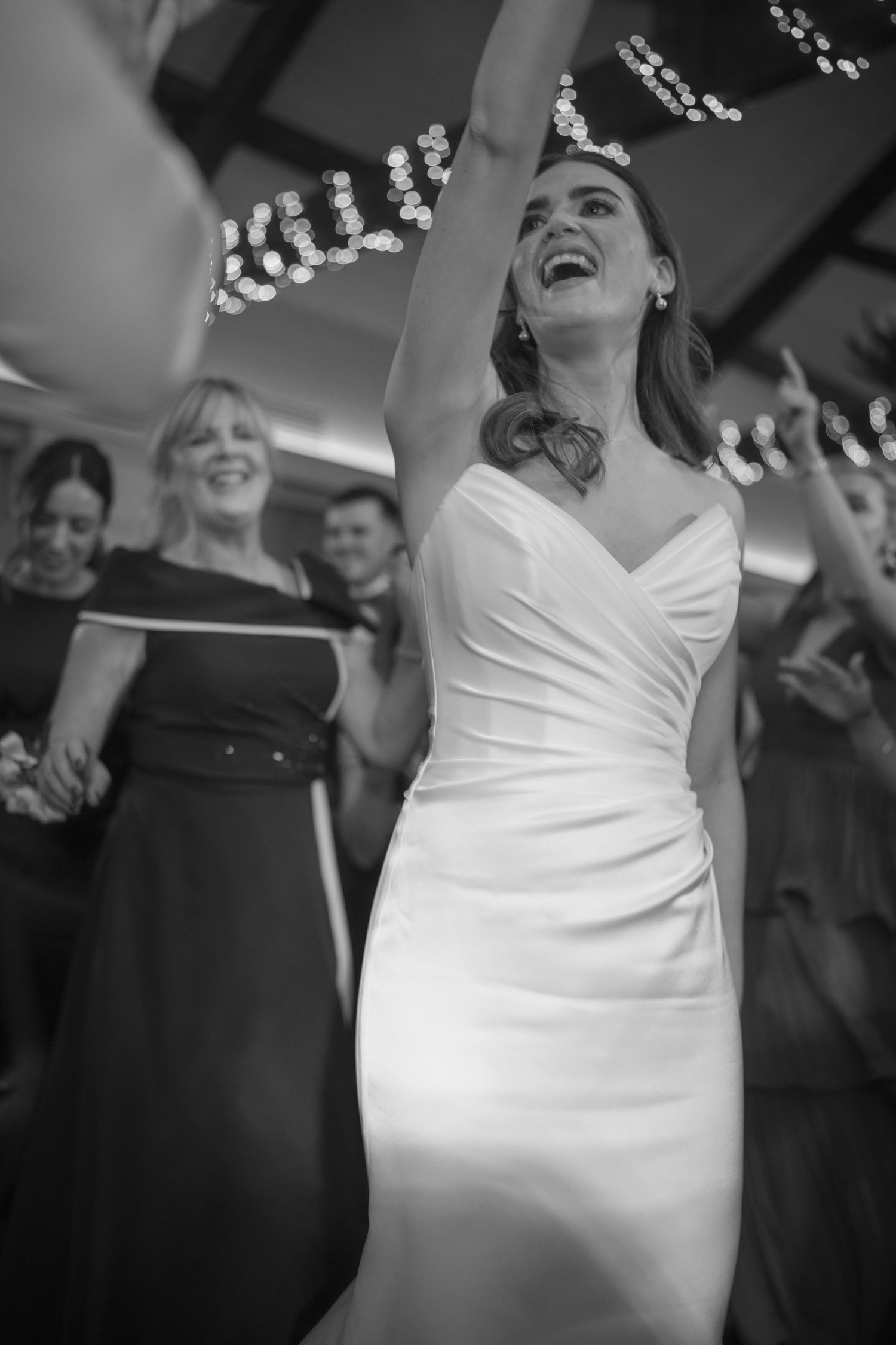 Bride in white wedding gown joyfully dancing with arms raised surrounded by guests at reception.