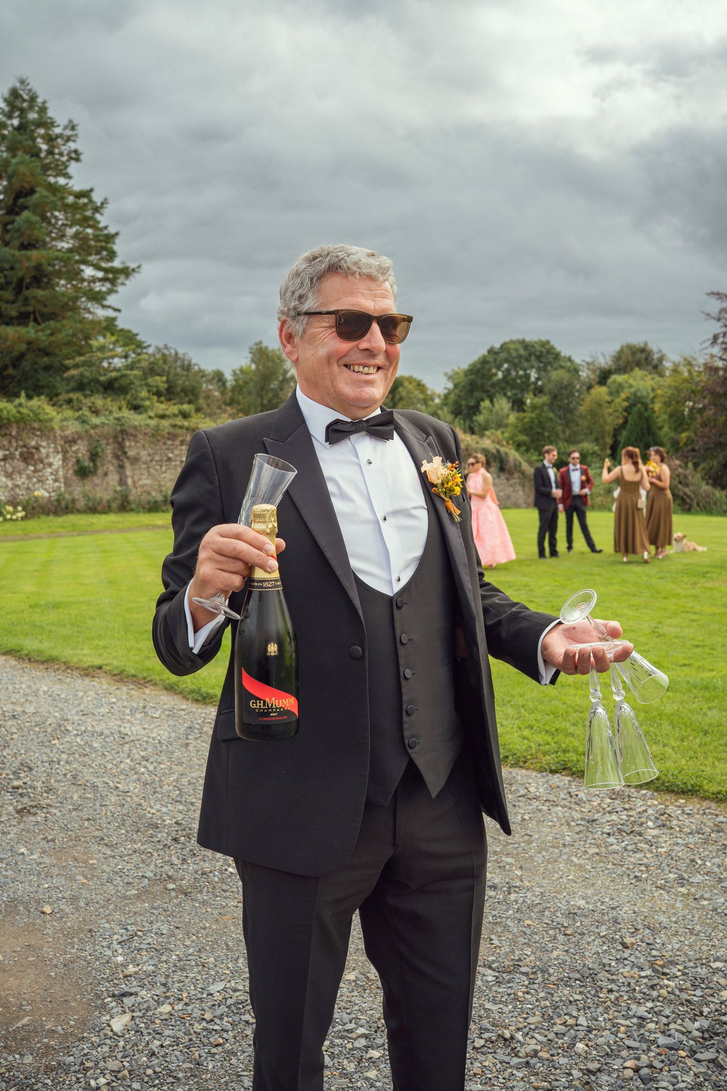 Father of the bride in tuxedo holding champagne bottle and glasses outdoors outdoor garden wedding reception in Co Meath.