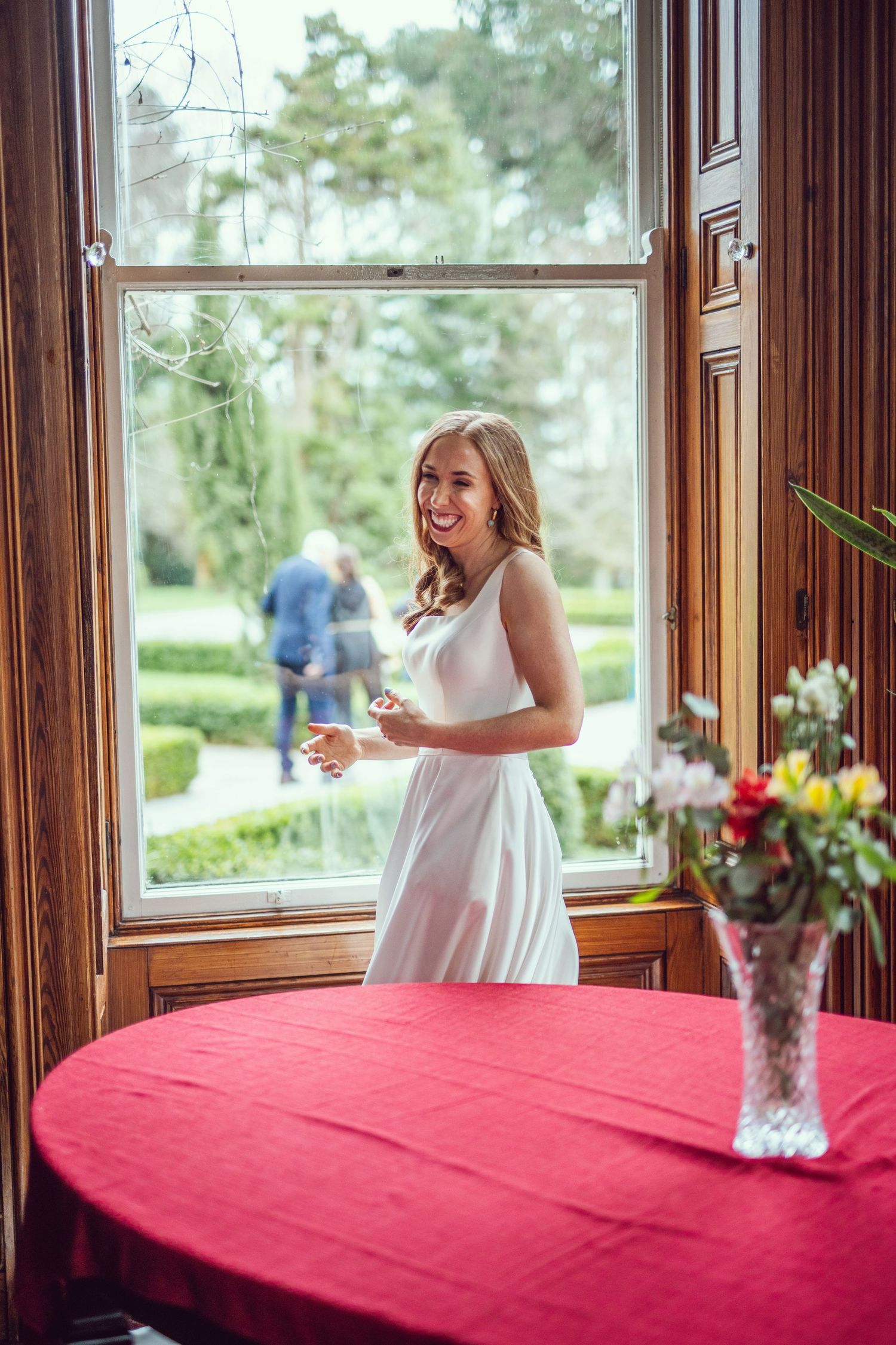 Smiling woman in white dress standing by large window with garden view indoors.