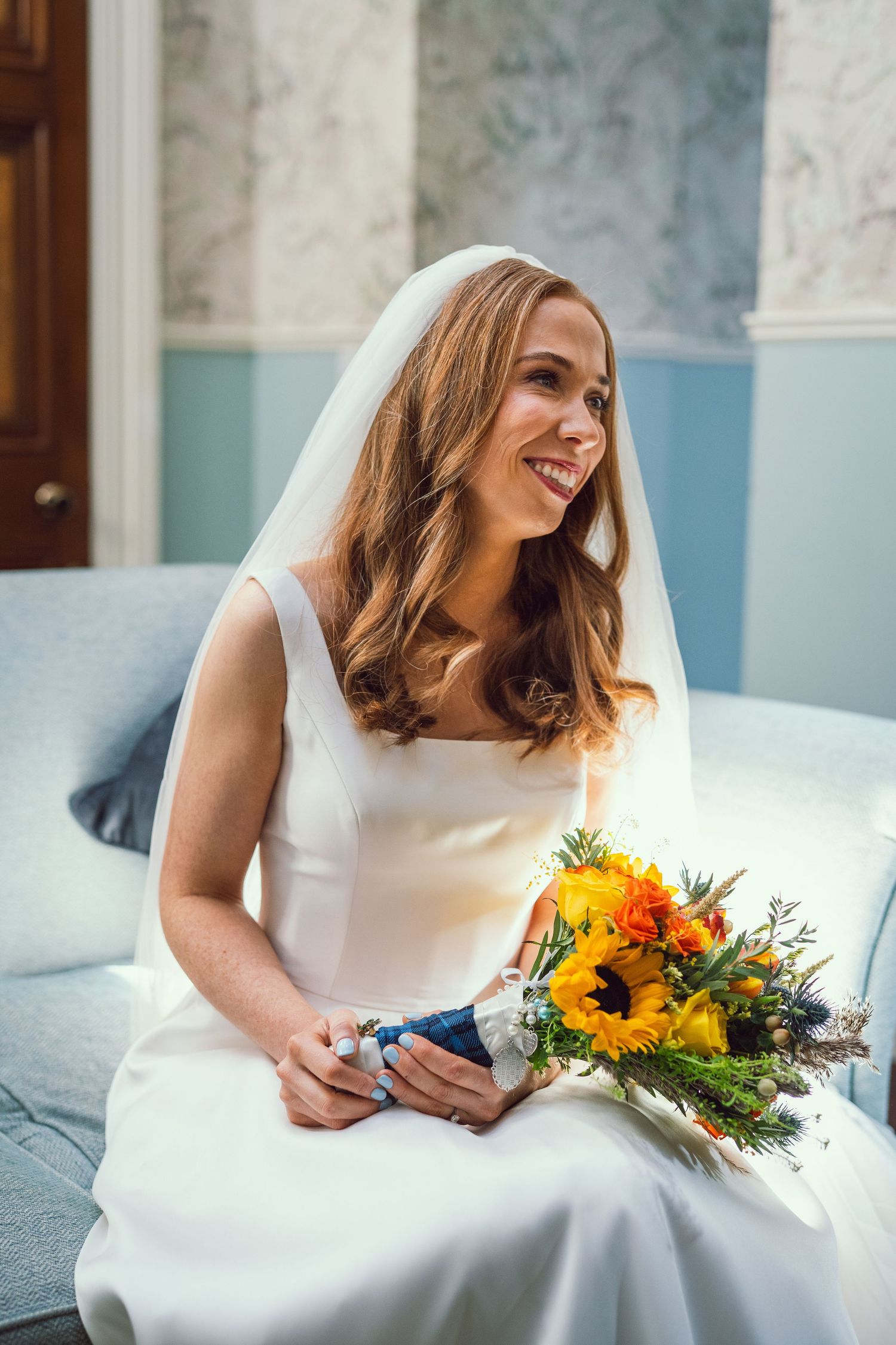 Smiling bride in white gown holding vibrant sunflower bouquet while seated indoors.