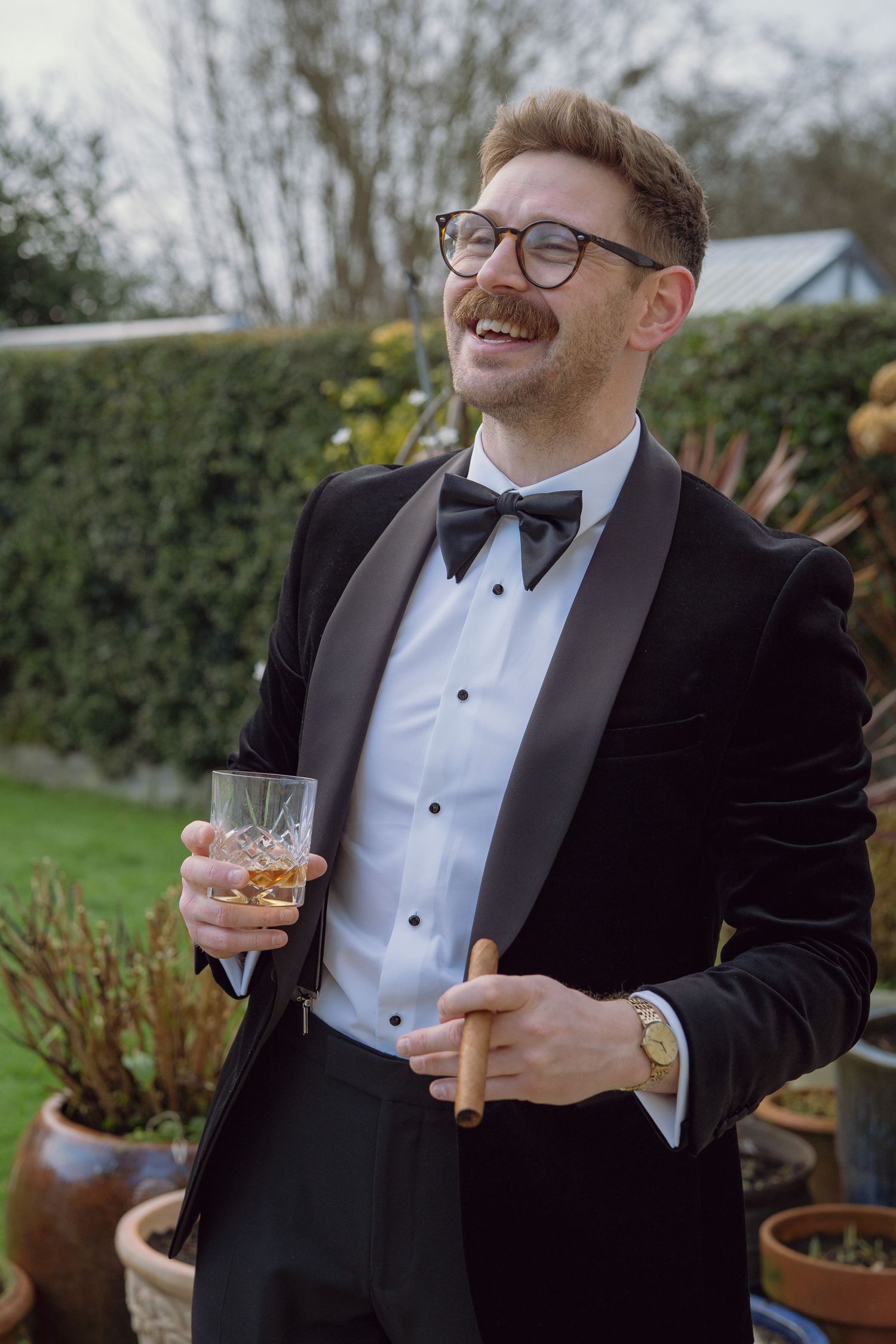 Smiling groom in black tuxedo with bow tie holds whiskey glass and cigar outdoors at wedding celebration.