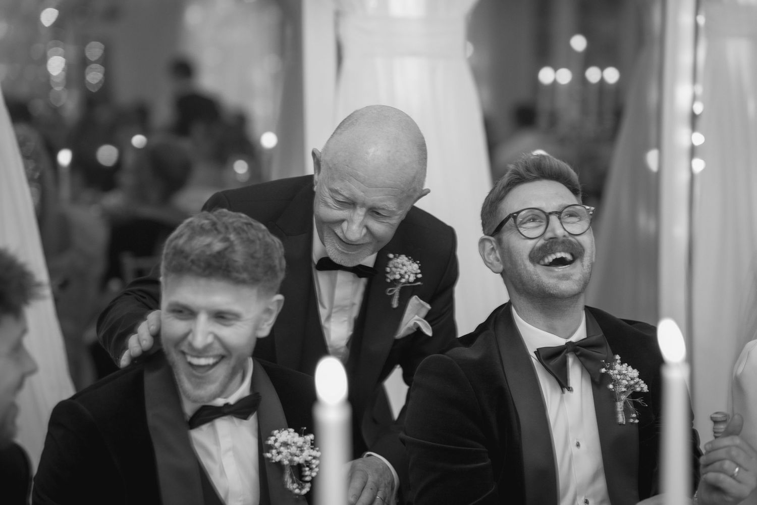 Three men in tuxedos sharing a joyful laugh at a wedding reception in black and white.
