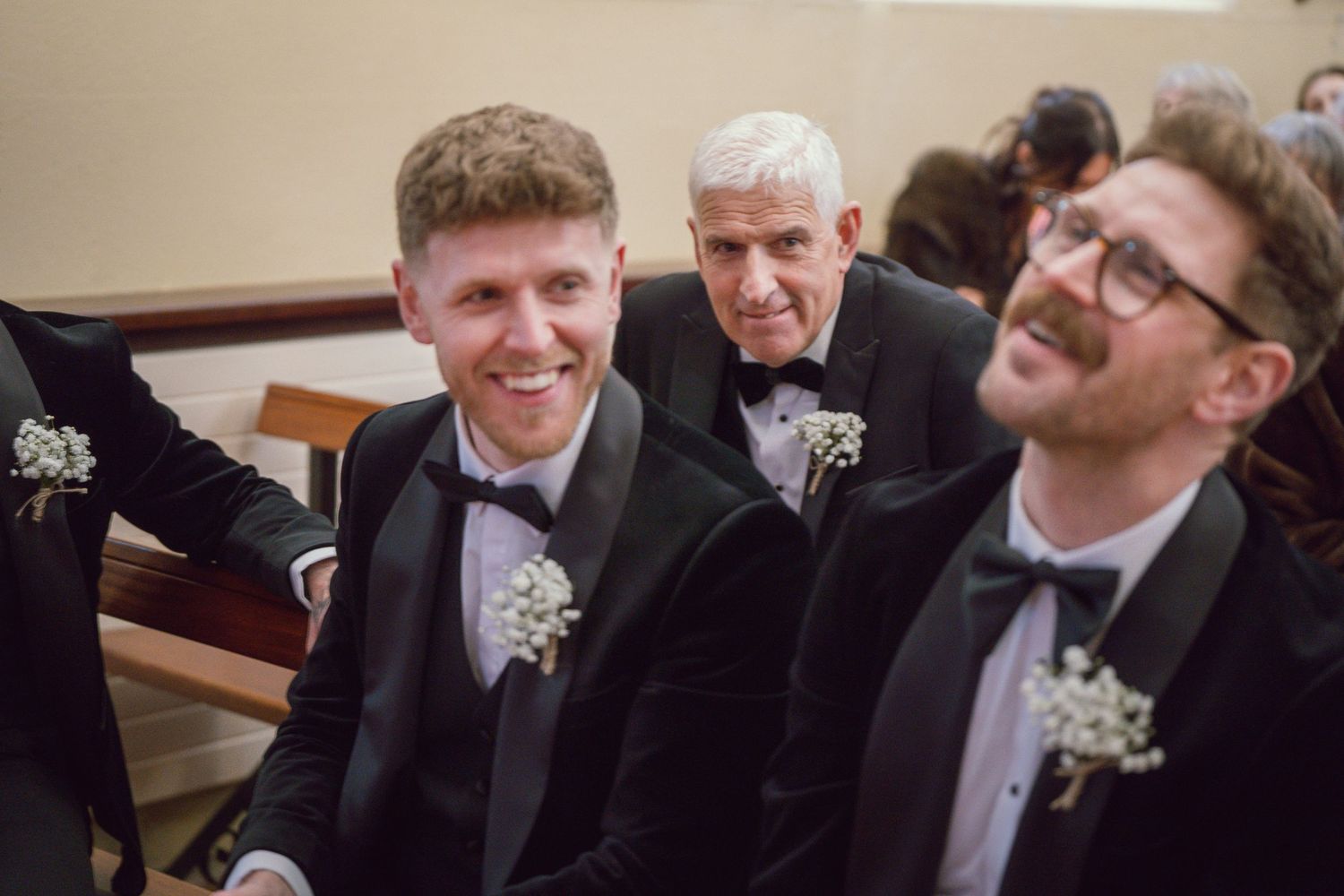 Groomsmen in black tuxedos smile and laugh together during a wedding ceremony.