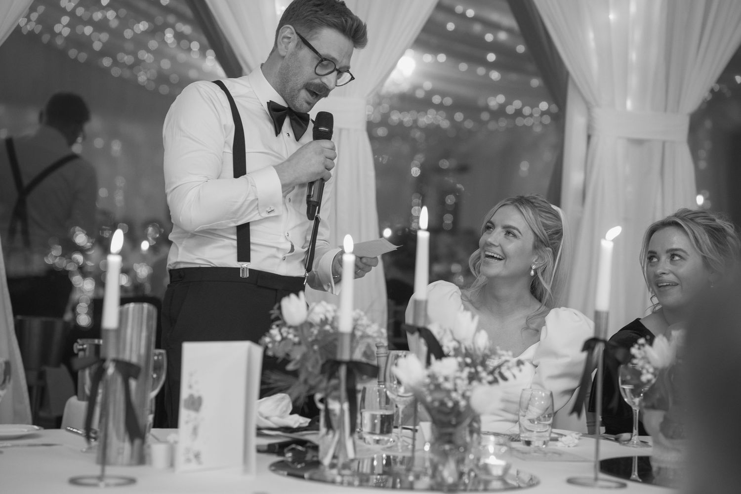 Groom speaking into microphone at wedding reception while bride smiles, black and white photo.