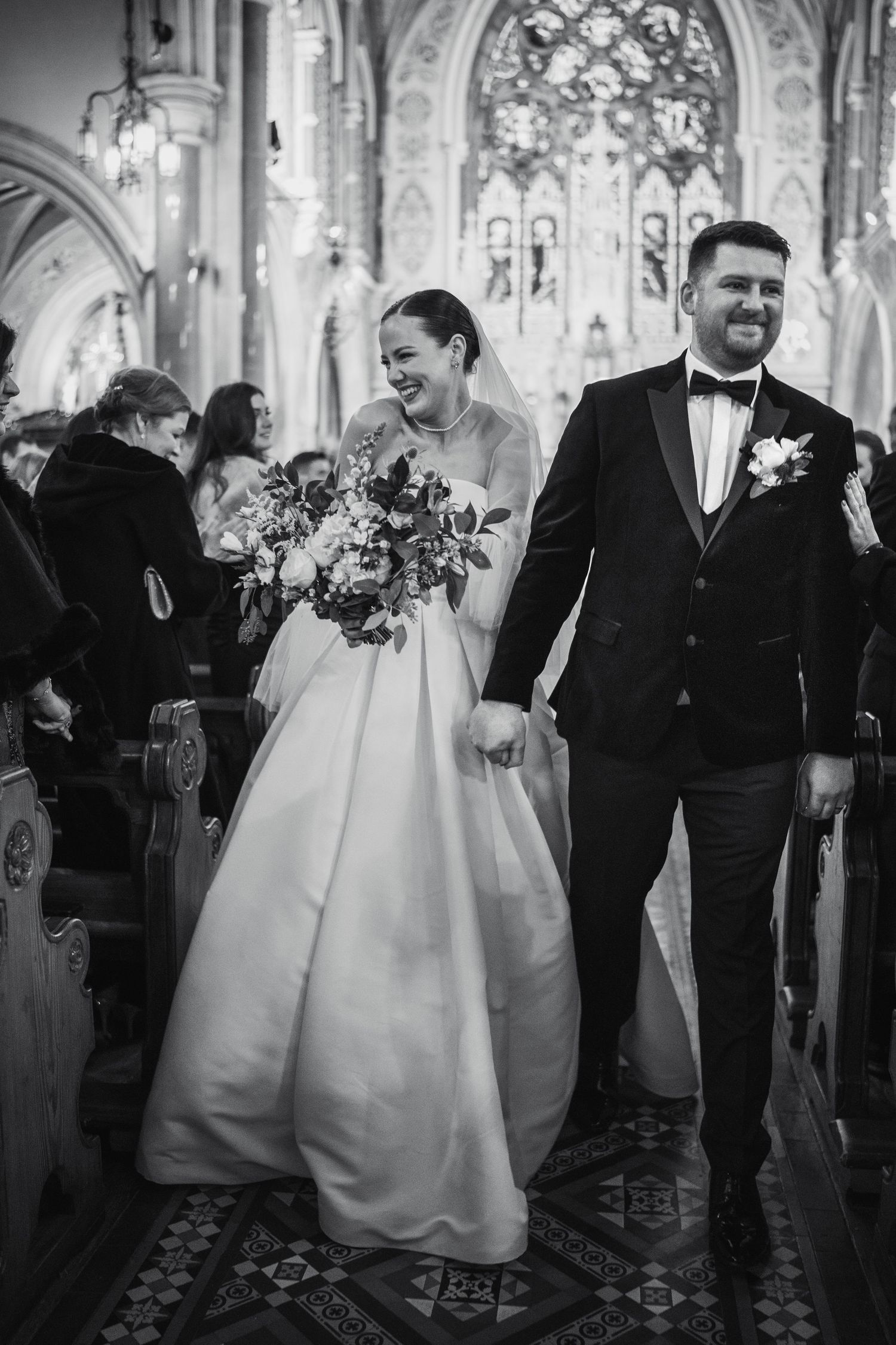 Bride and groom walk down church aisle laughing, bride holds floral bouquet, groom in tuxedo, black and white wedding photo.