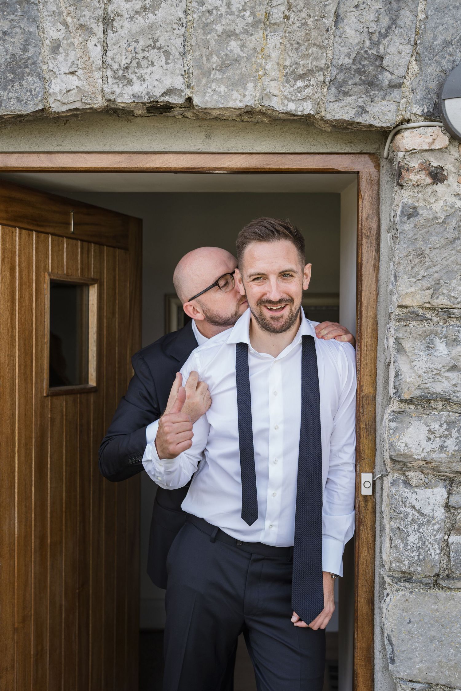 Bald groomsman playfully kissing the groom on the cheek in a rustic stone doorway.