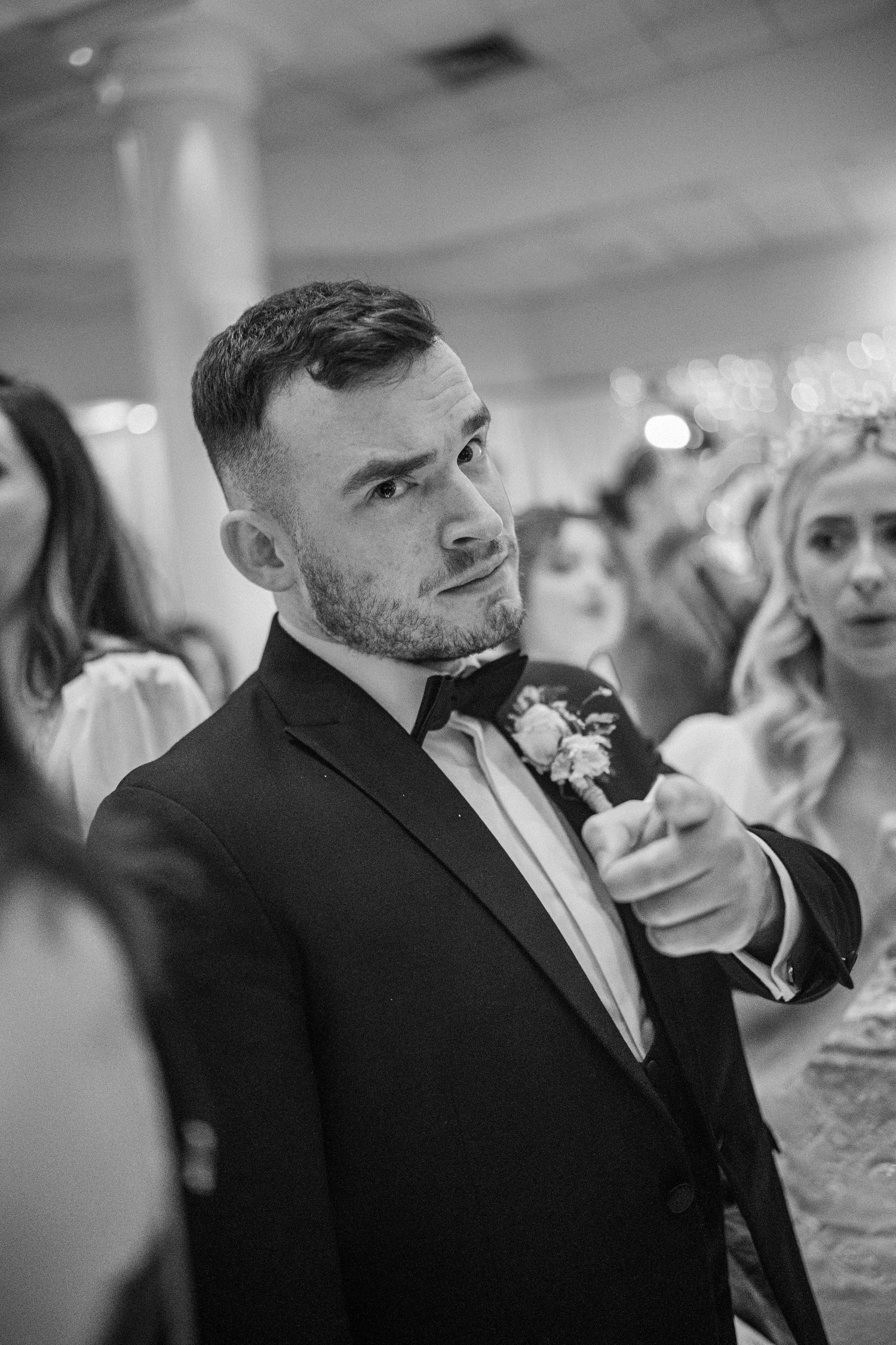Dapper groomsman in black tuxedo with boutonniere pointing at camera during wedding reception.