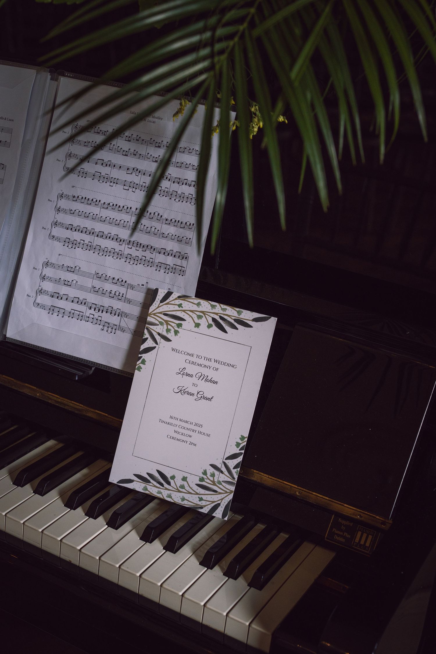 Wedding program resting on piano keys beside sheet music and tropical palm leaves.