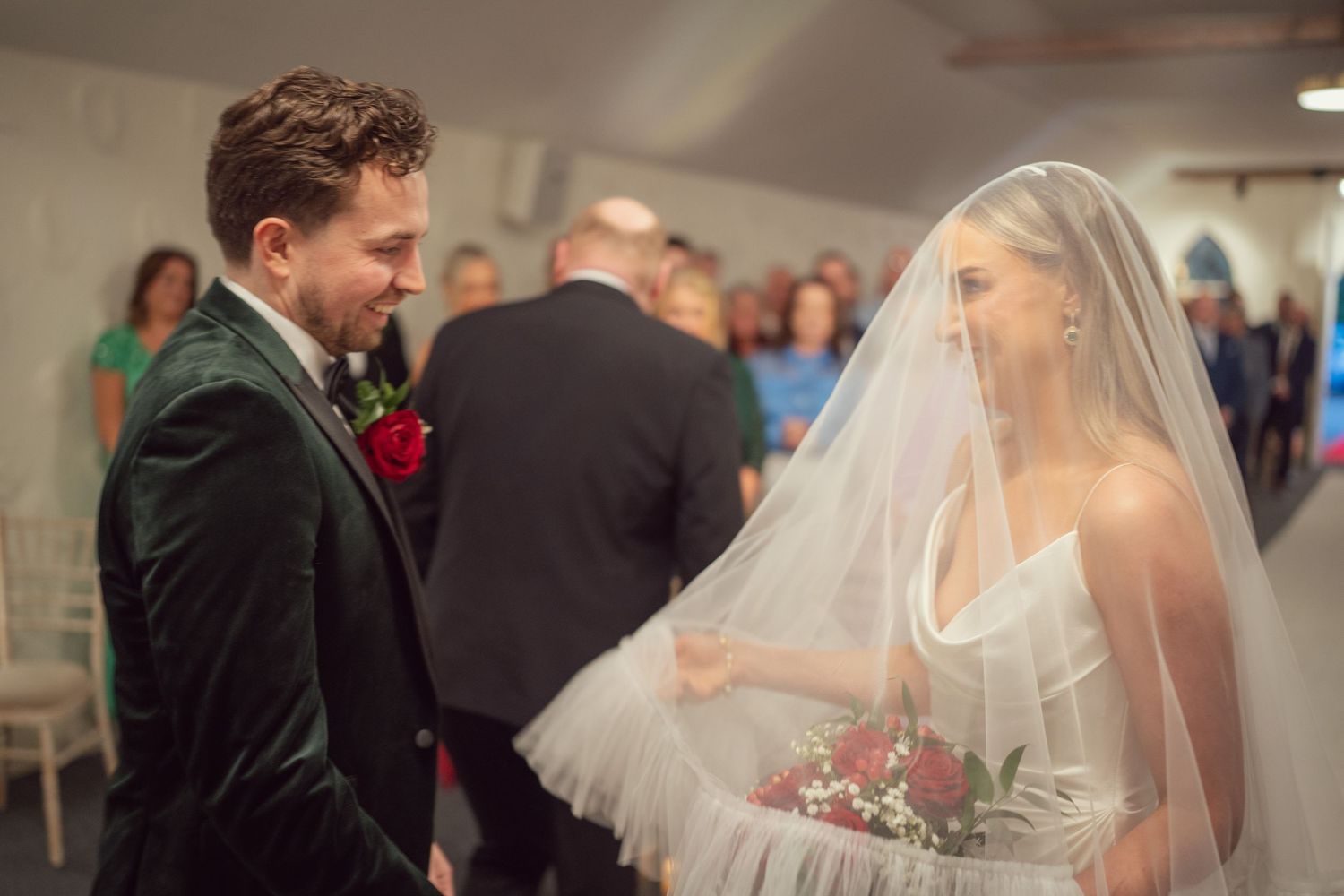 Groom in green velvet suit lifting bride's veil at wedding ceremony altar smiling.